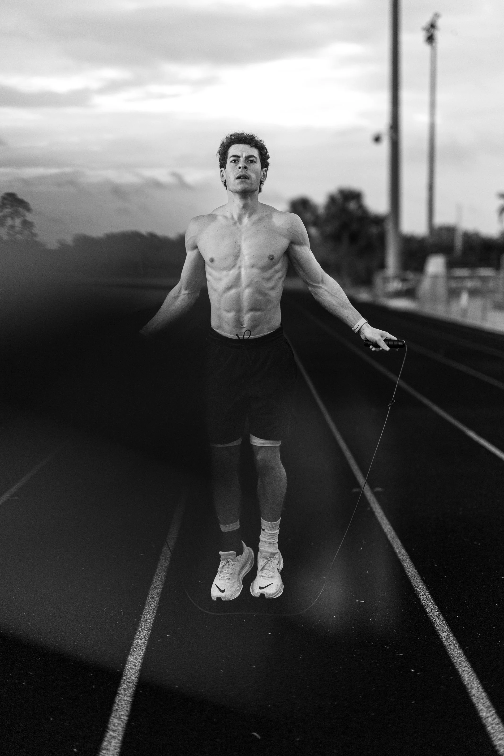 Black and white photo of a shirtless man jogging on an outdoor track, holding a jump rope, with a cloudy sky and trees in the background.