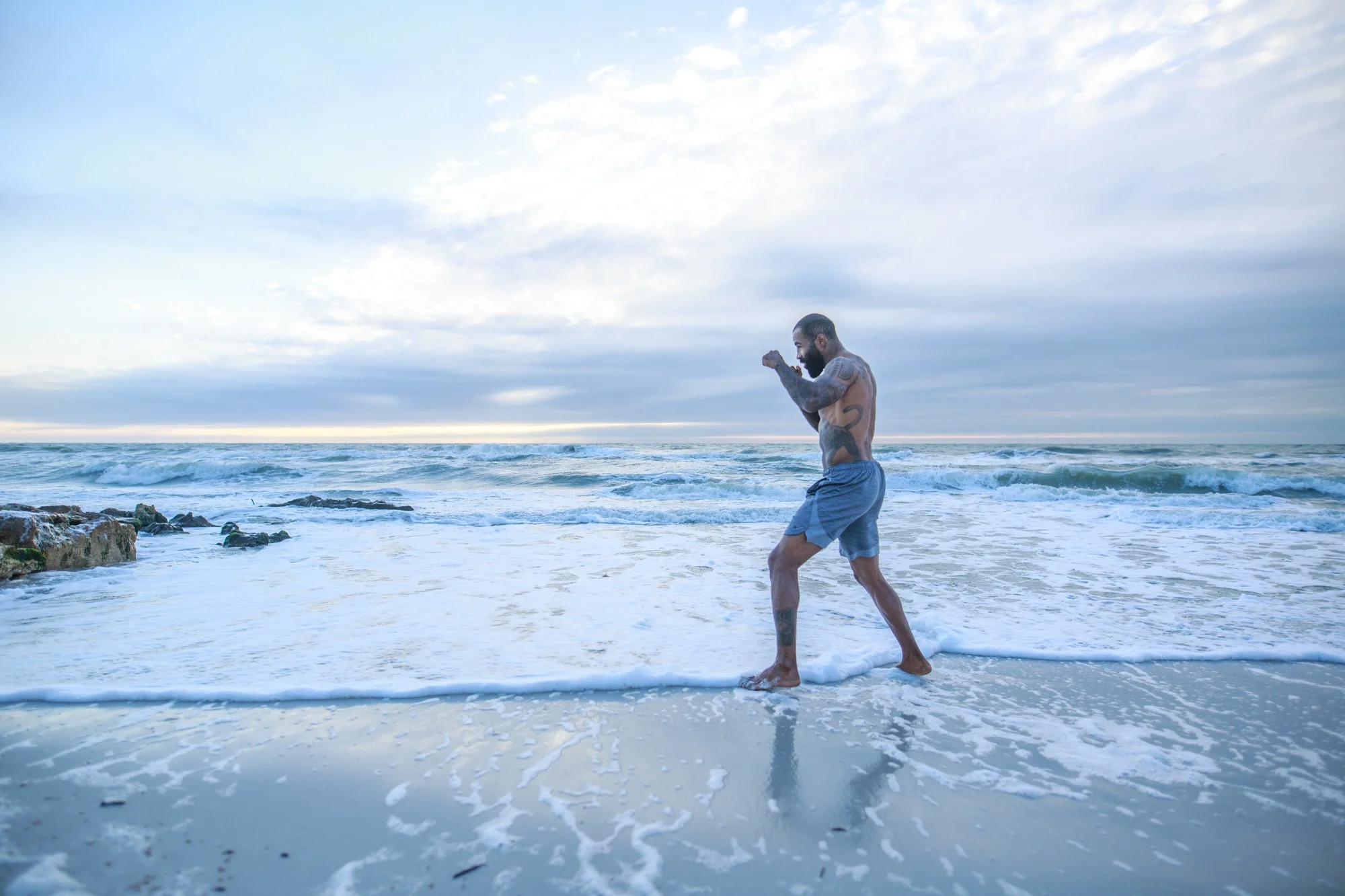 A man with tattoos practicing boxing on the beach at sunset, standing in the ocean with waves around his feet. Cosmo Alexandre, muay thai