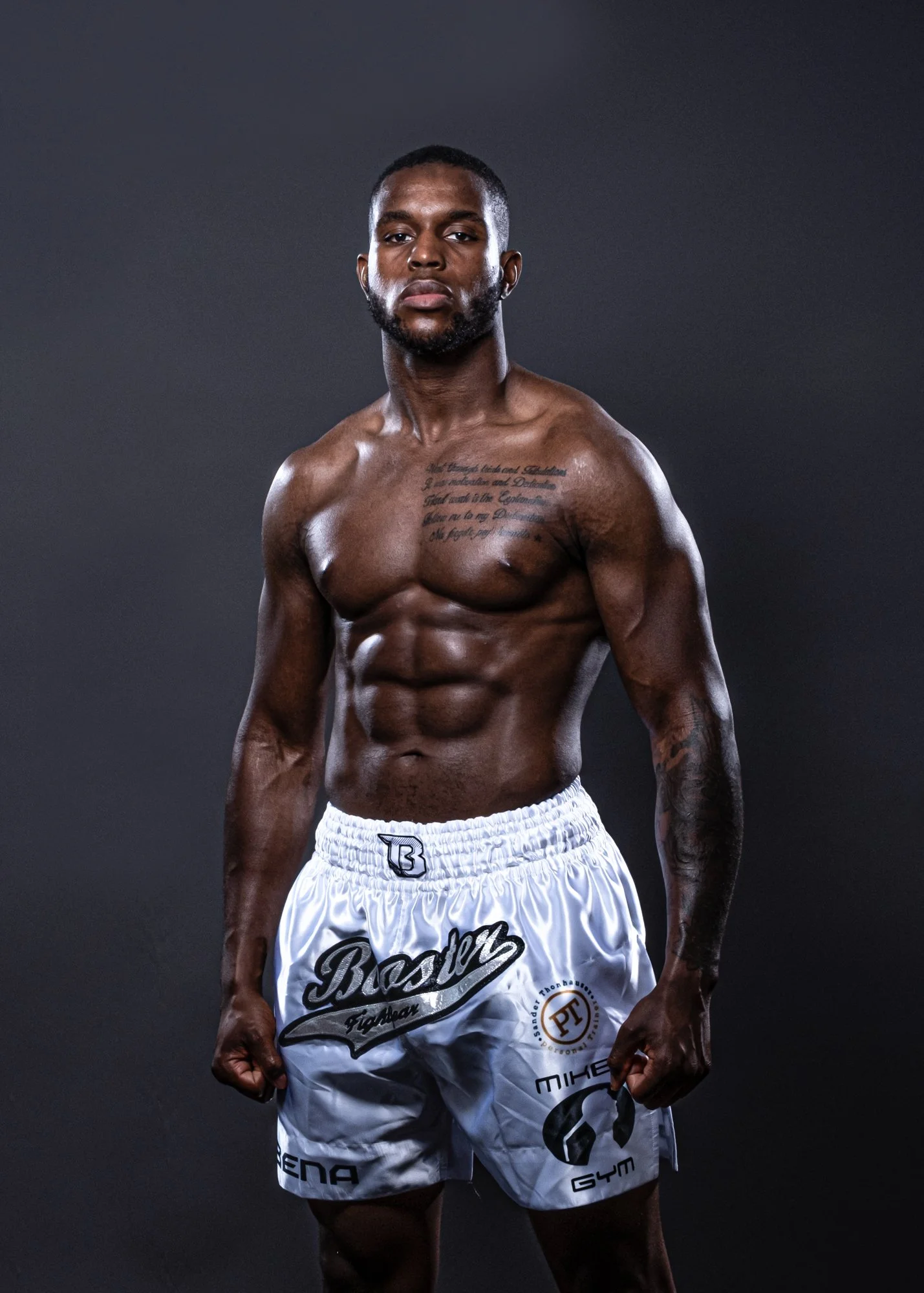 A muscular Black man standing shirtless with white boxing shorts against a dark background.