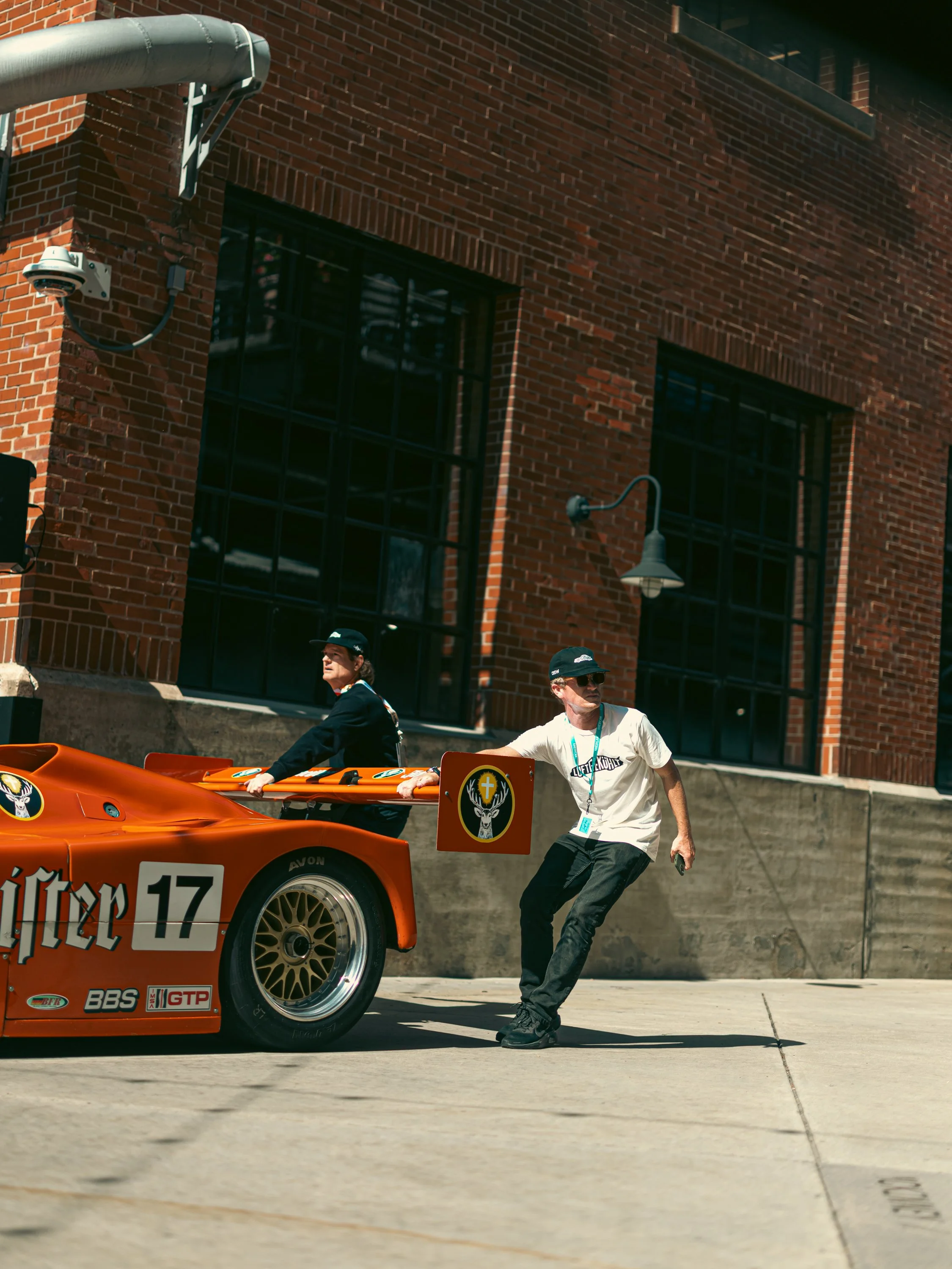 Two men are standing beside an orange race car with sponsorship stickers, in front of a brick building with black-framed windows. One man is wearing a white T-shirt, black pants, and a black cap, and the other man is wearing a black jacket and black 