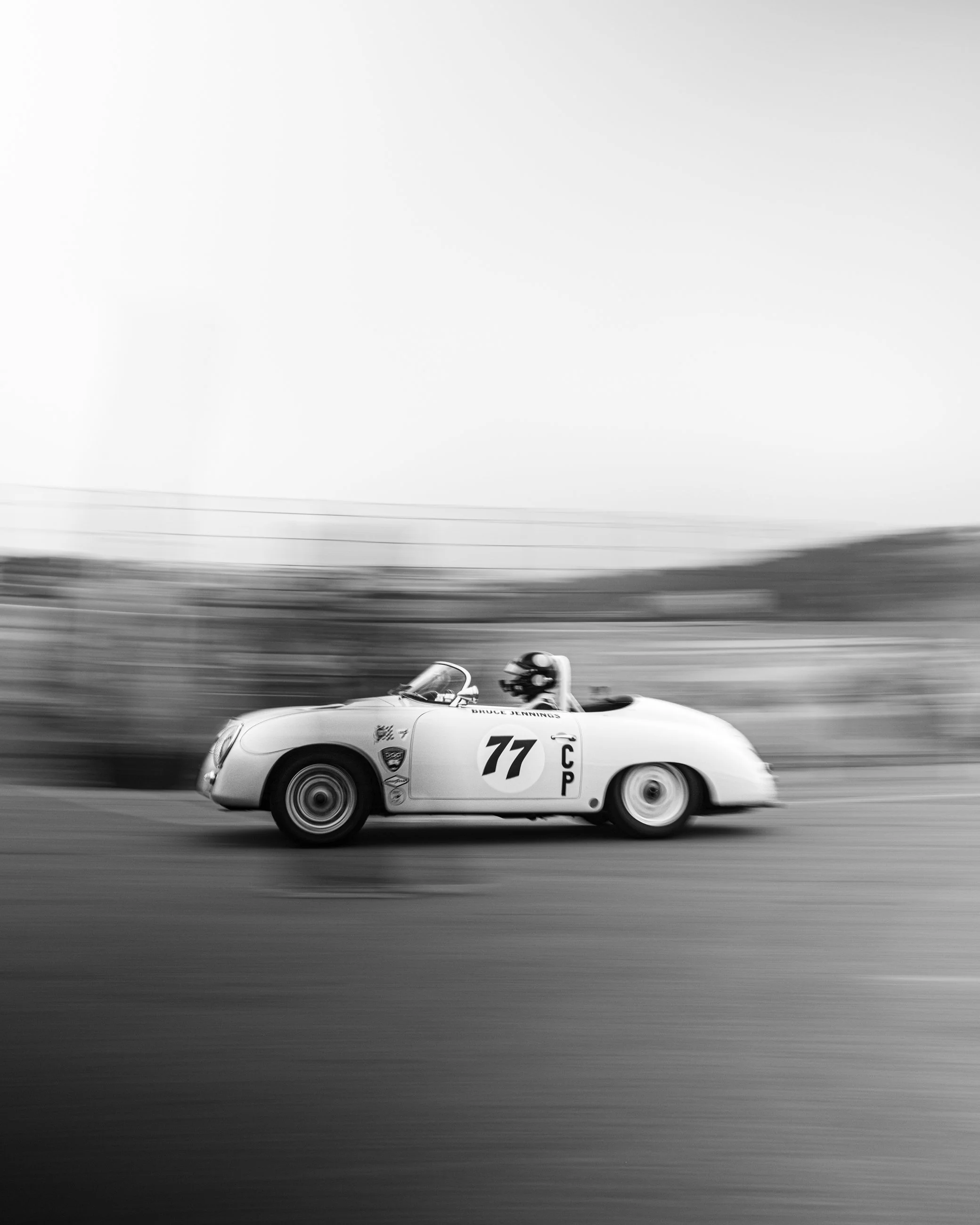 Black and white photograph of a vintage race car on the track, with visible number 77 on the side, passing by in motion, with blurred background indicating speed.