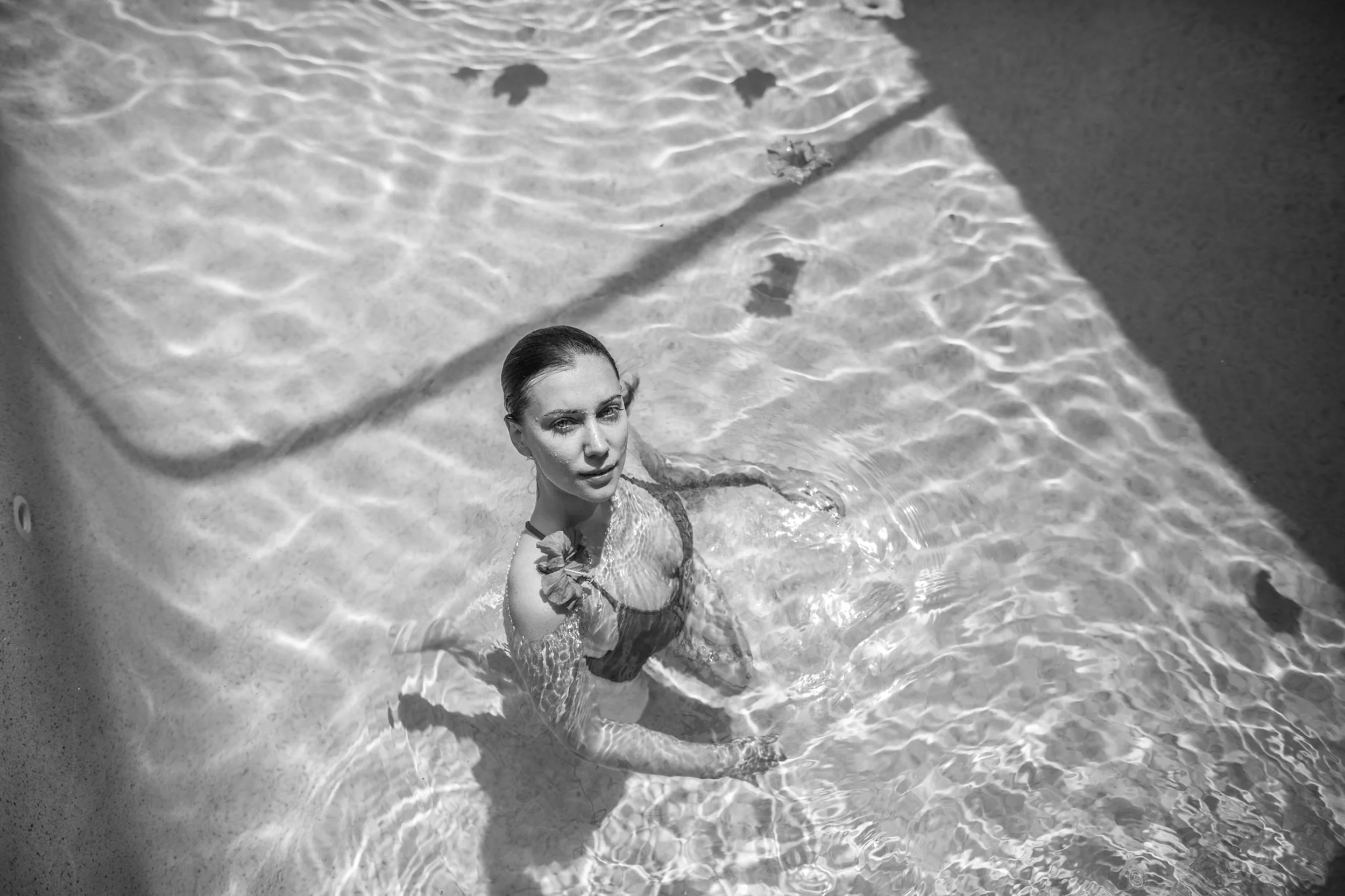 A woman with wet hair in a swimsuit standing in a pool with sunlight reflecting on the water's surface.