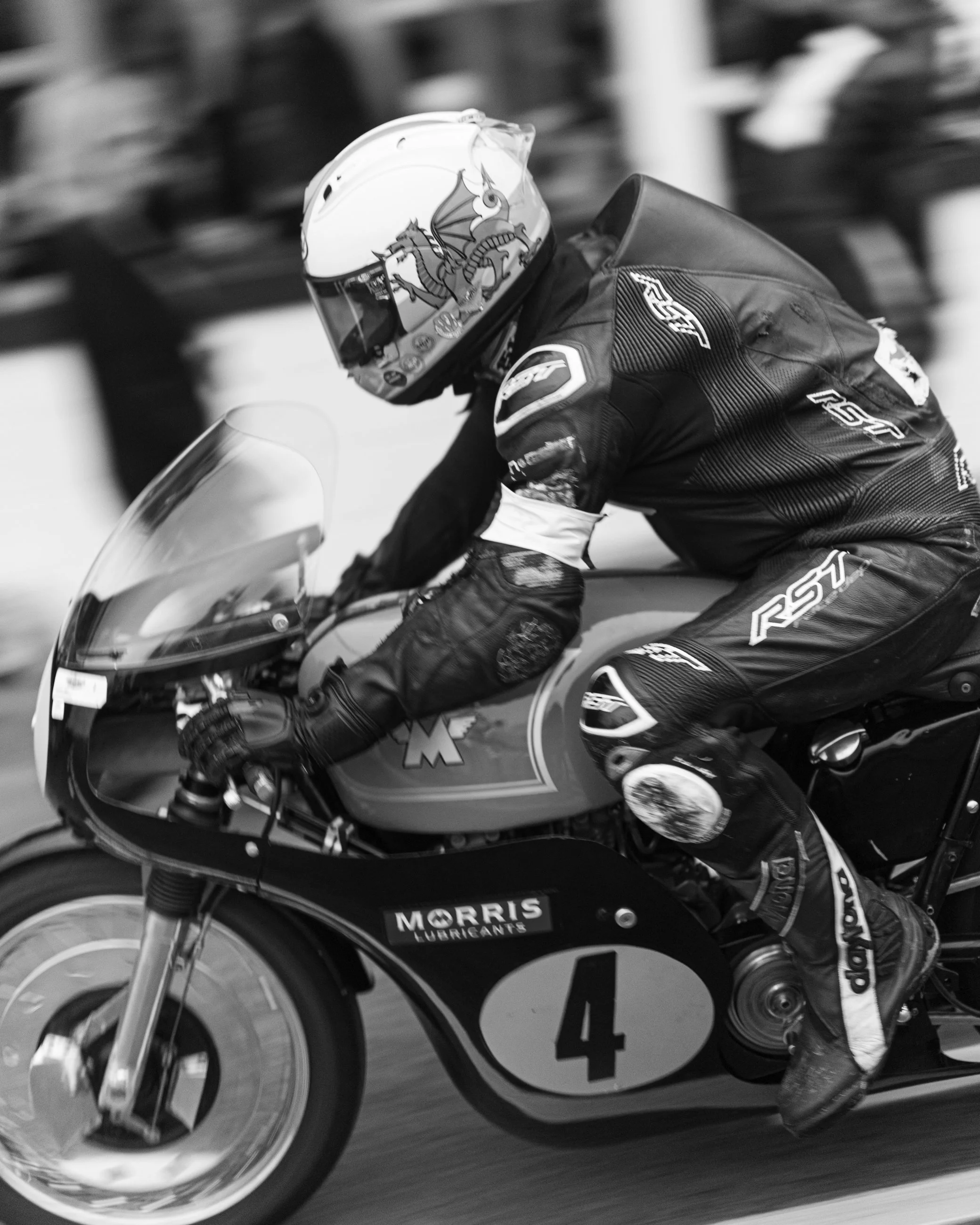 Close-up of a motorcycle racer in black racing suit with white accents, riding a racing motorcycle on a track, wearing a helmet featuring a dragon design. Goodwood Revival