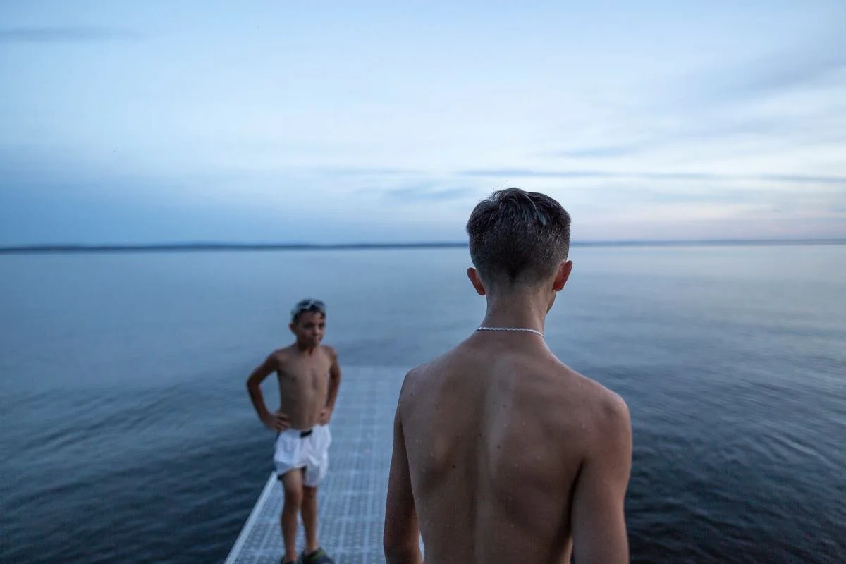 Two shirtless young boys on a dock by a calm body of water, with one boy facing away from the camera and the other standing further back smiling.