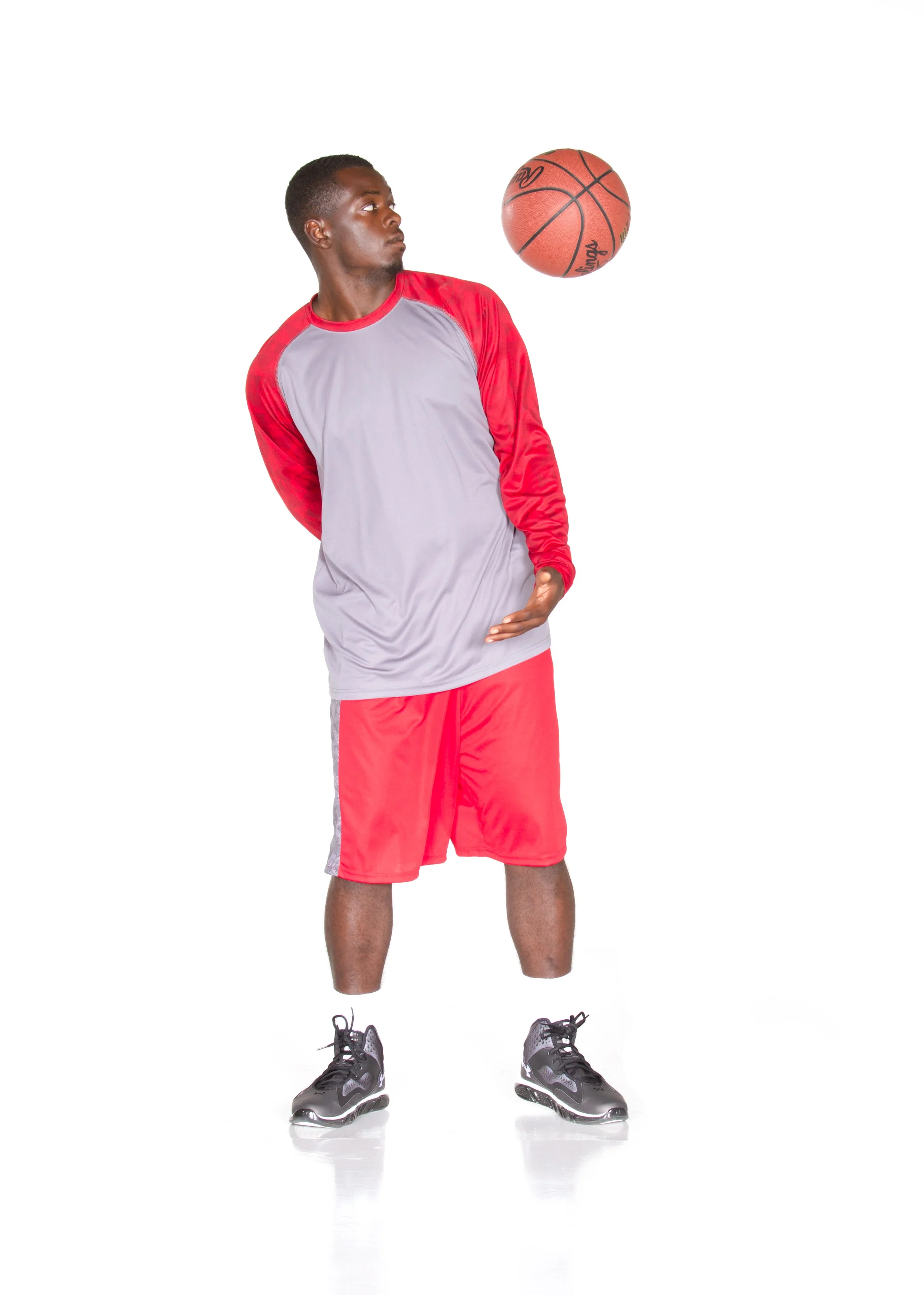A young man standing against a white background, wearing red shorts and a gray and red sports shirt, with black athletic shoes, is in the process of dribbling a basketball.