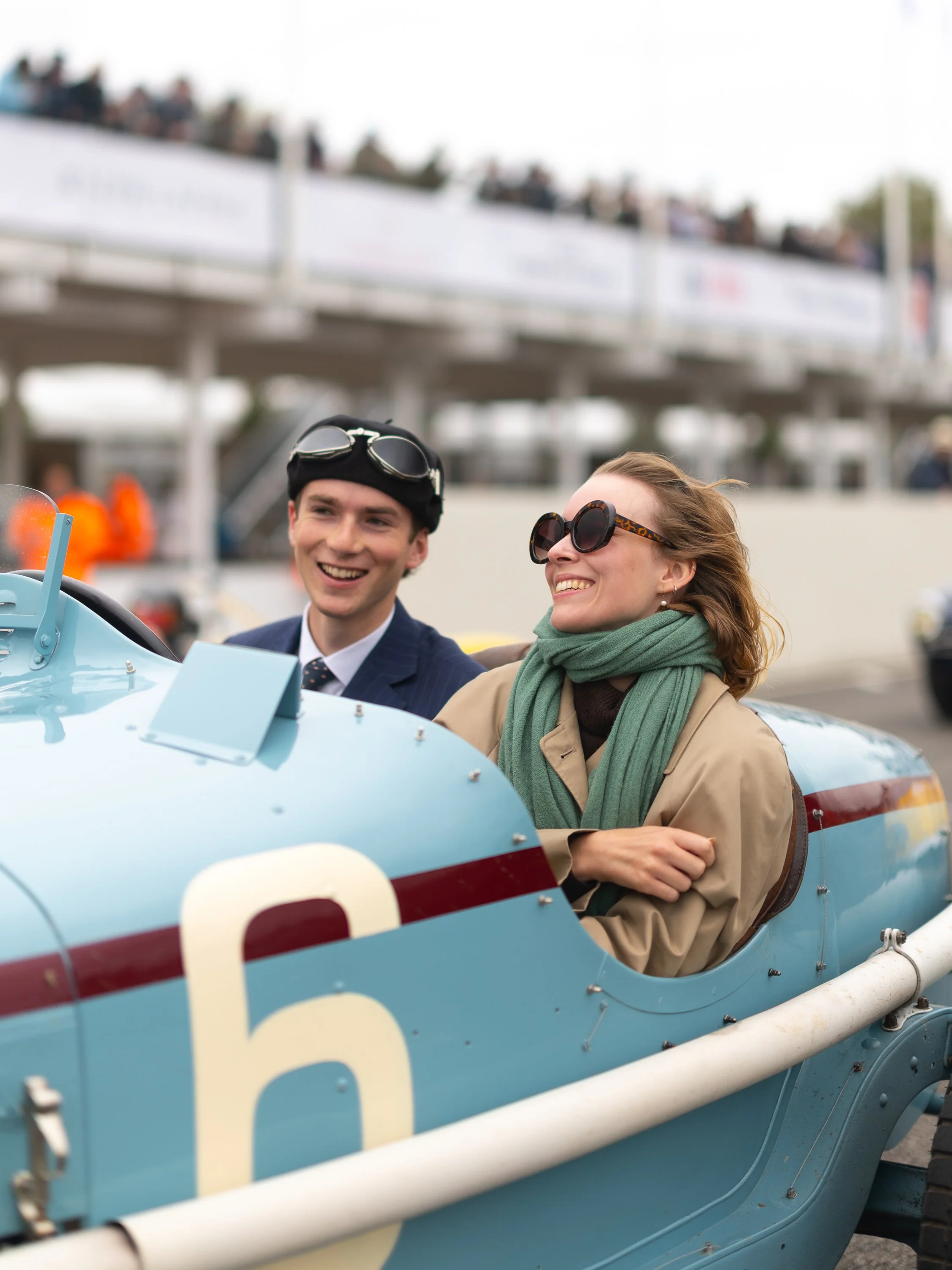 A vintage race car with a blue body and white racing number, driven by a man and a woman, both wearing goggles and smiling, at a racing event with spectators and a grandstand in the background. Goodwood Revival.