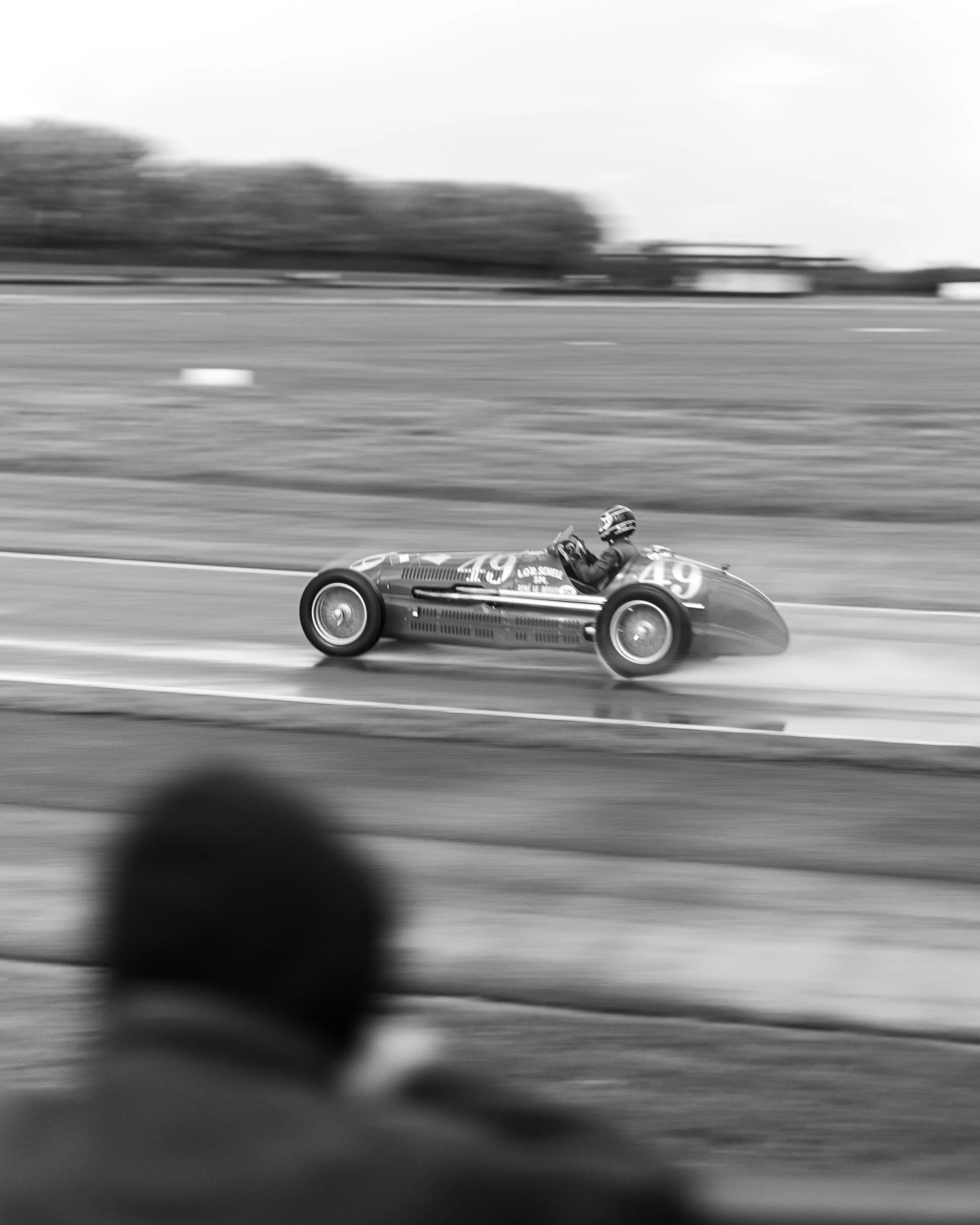 A vintage race car with a driver in a helmet speeding down a racetrack, motion blurred background in black and white.
