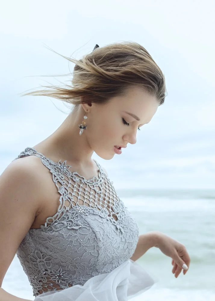 A young woman with long hair blowing in the wind, wearing a gray lace dress and earrings, standing near the ocean.
