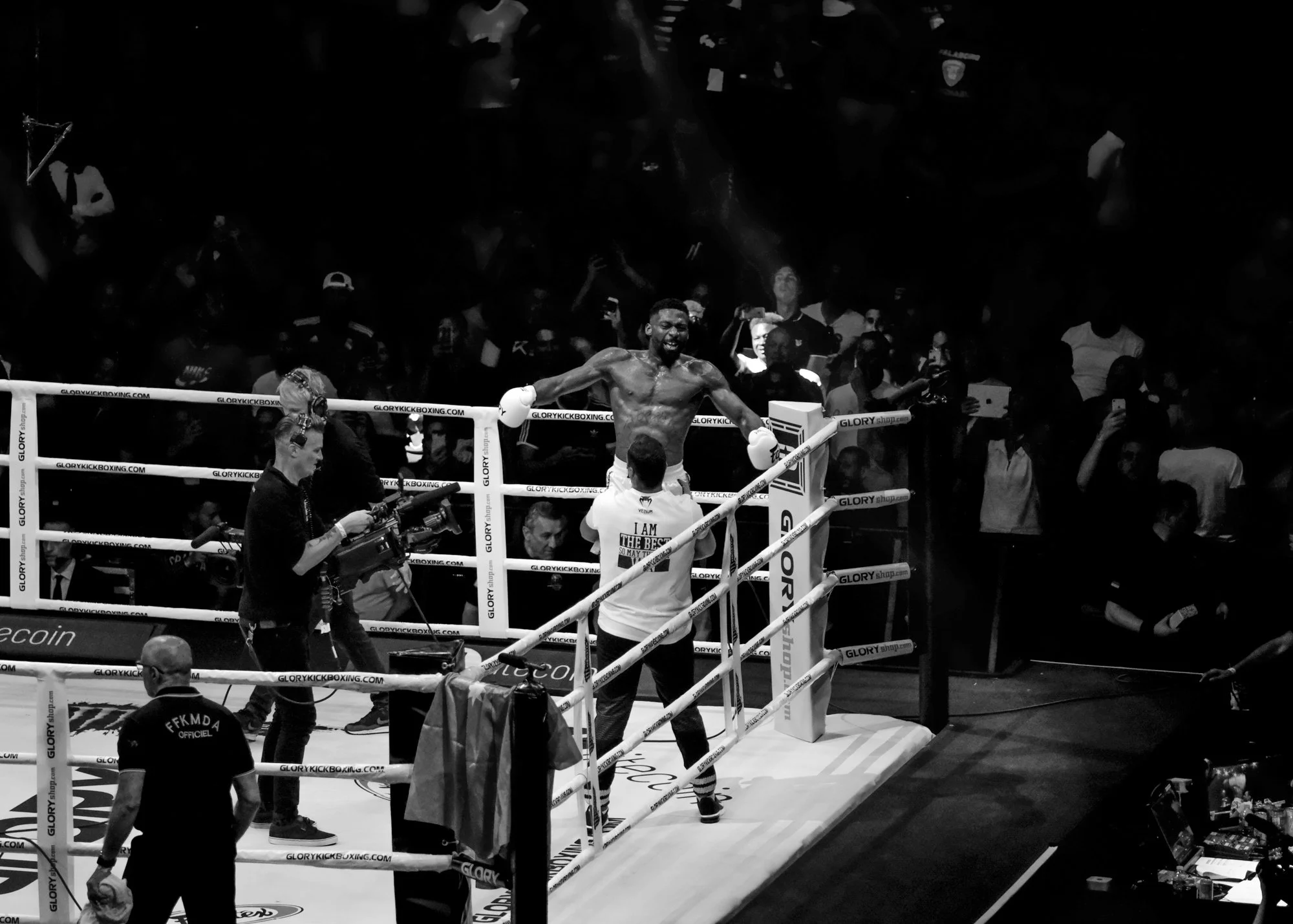 A victorious male kickboxer celebrates in the boxing ring with a big smile, surrounded by officials and audience members, during a match inside a sports arena.