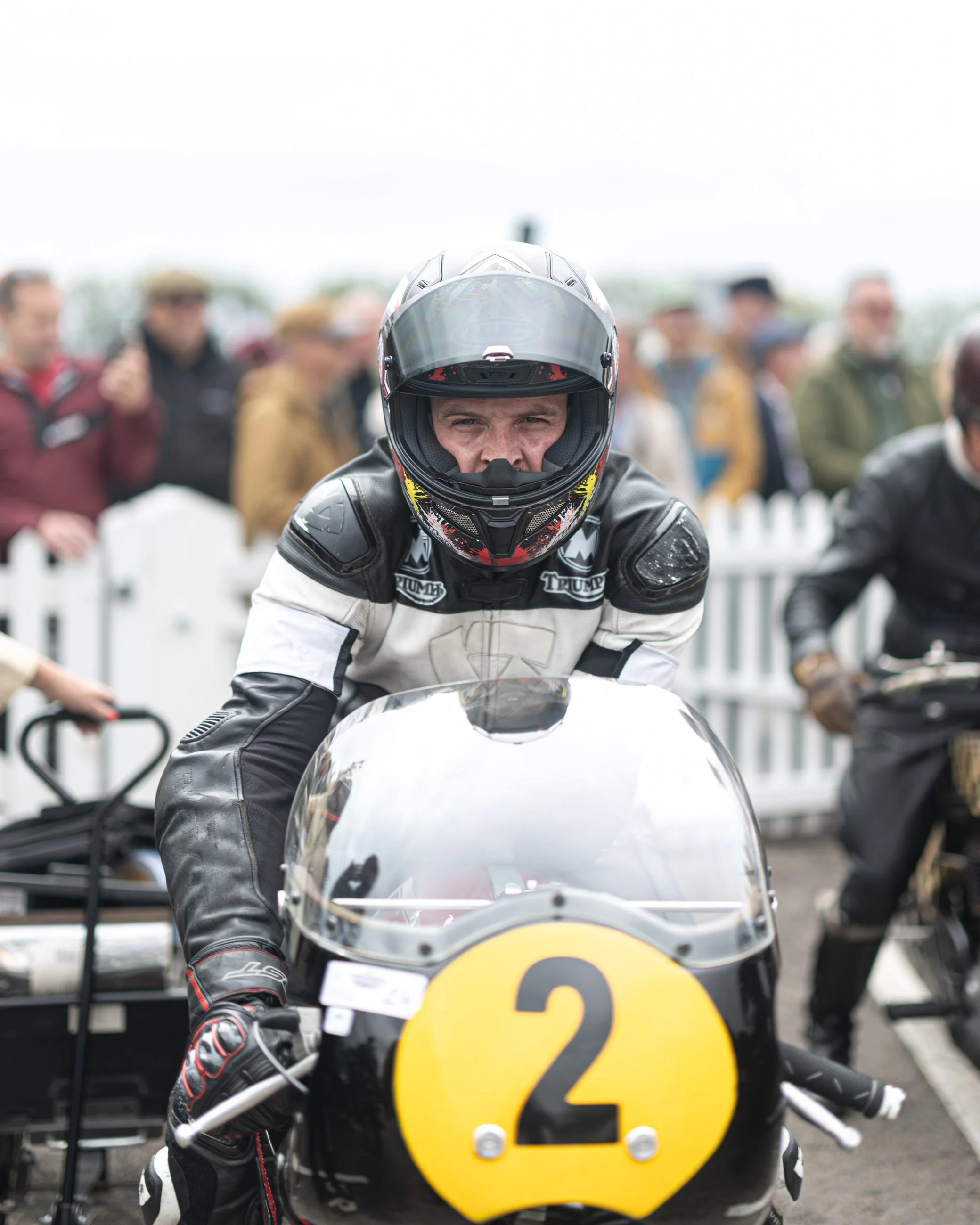 Close-up of a motorcycle racer wearing a black and white racing suit and helmet, sitting on a motorcycle with the number 2 on it, at a racing event with spectators in the background.