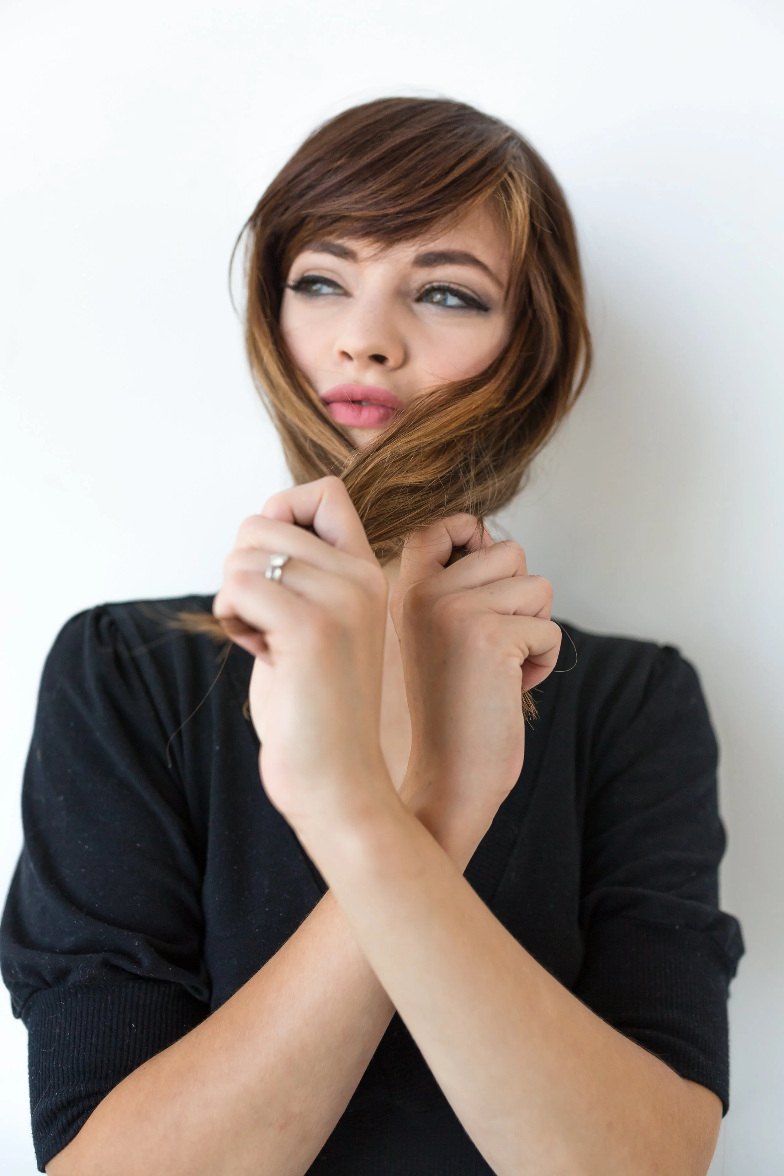 A woman with light skin and reddish-brown hair is holding a lock of her hair in front of her face, looking to the side with a thoughtful expression. She is wearing a black top.
