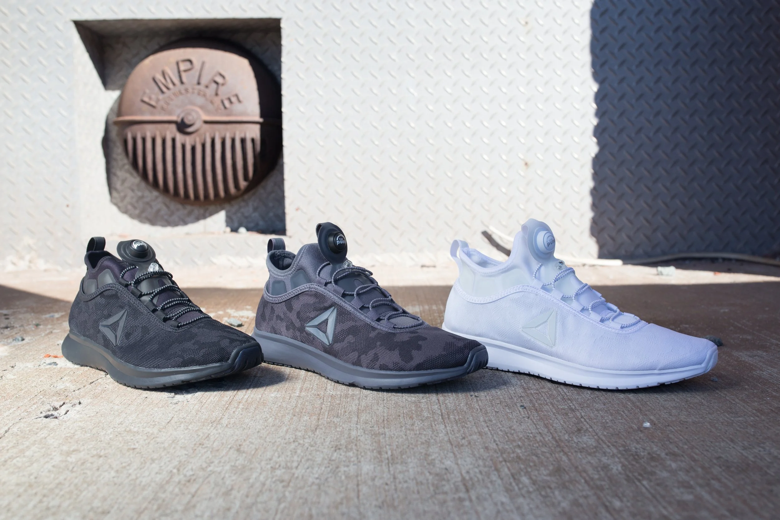 Three pairs of athletic shoes in black, gray, and white, placed on a textured surface in front of a metallic wall with a fire alarm and shadow. Reebok.