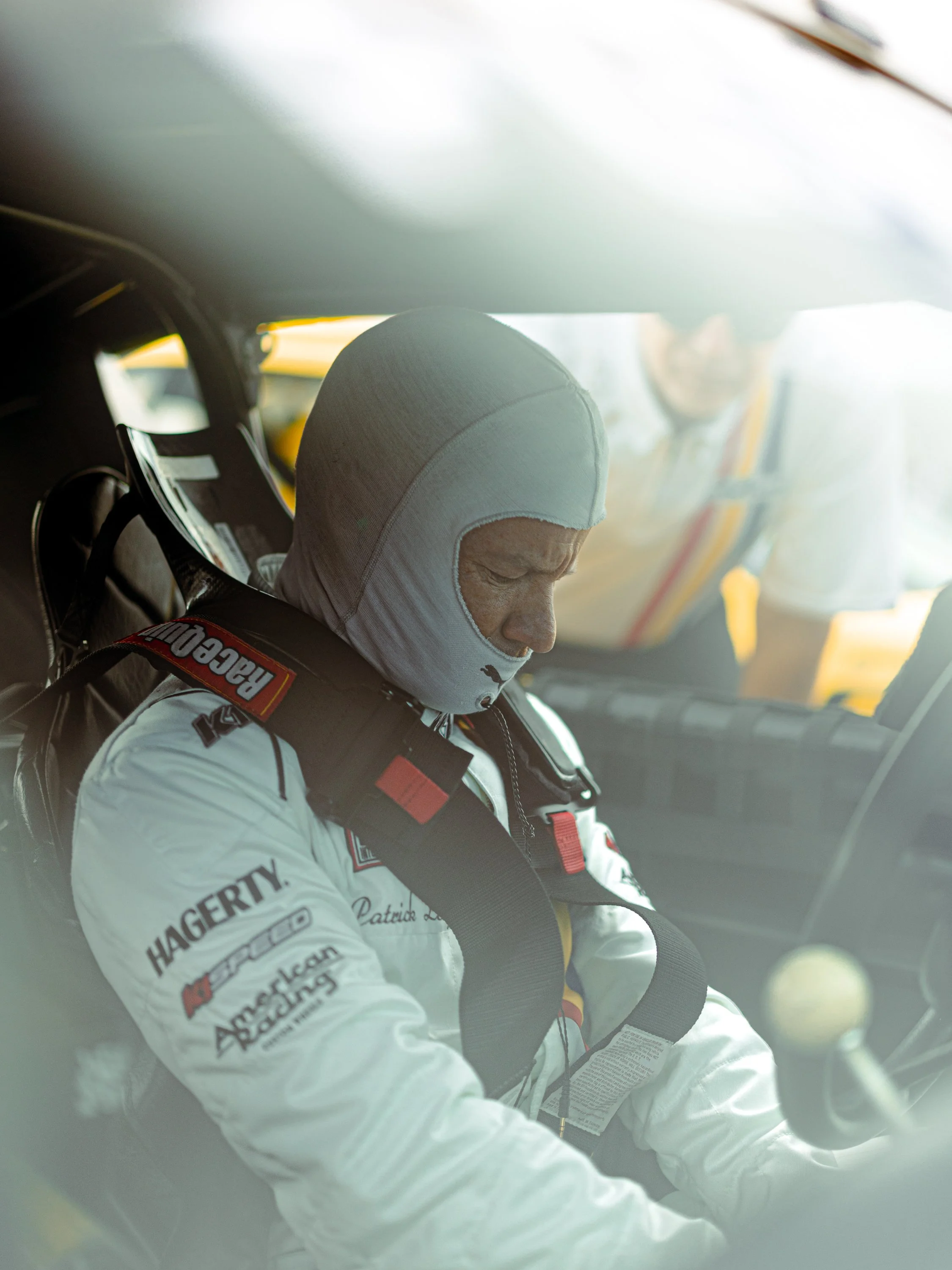 A race car driver wearing a white racing suit and a white hood is sitting in a car, preparing for a race. The race suit has sponsor logos and patches on it, and the driver appears to be focused. Patrick Long