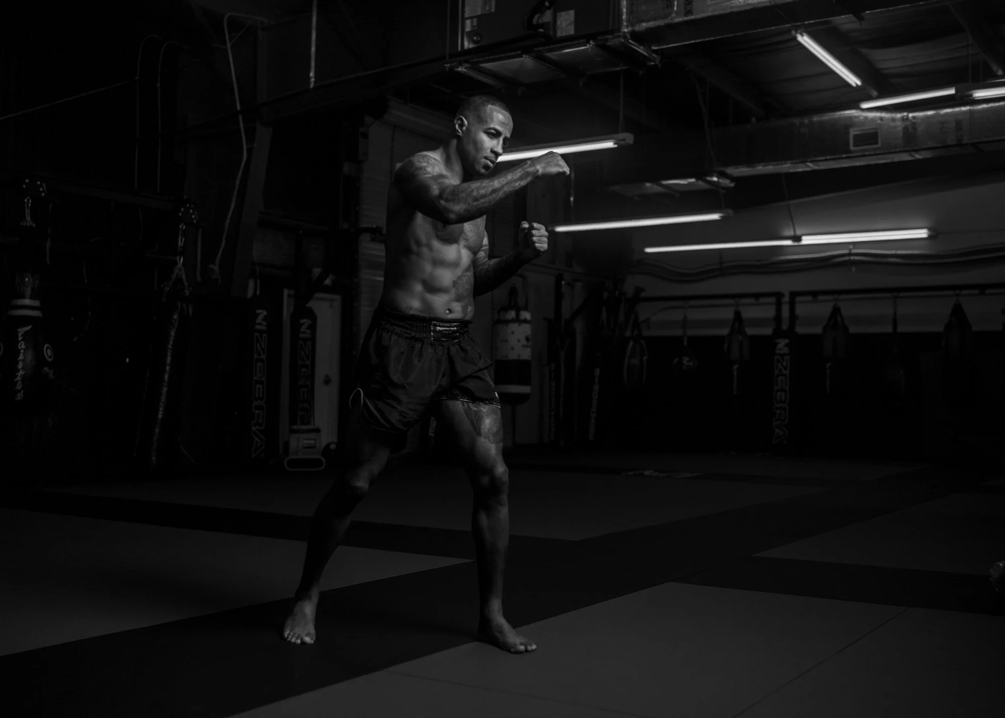 A shirtless man with tattoos practicing boxing or martial arts in a dark gym, standing in a fighting stance with fists raised. Cosmo Alexandre, muay thai