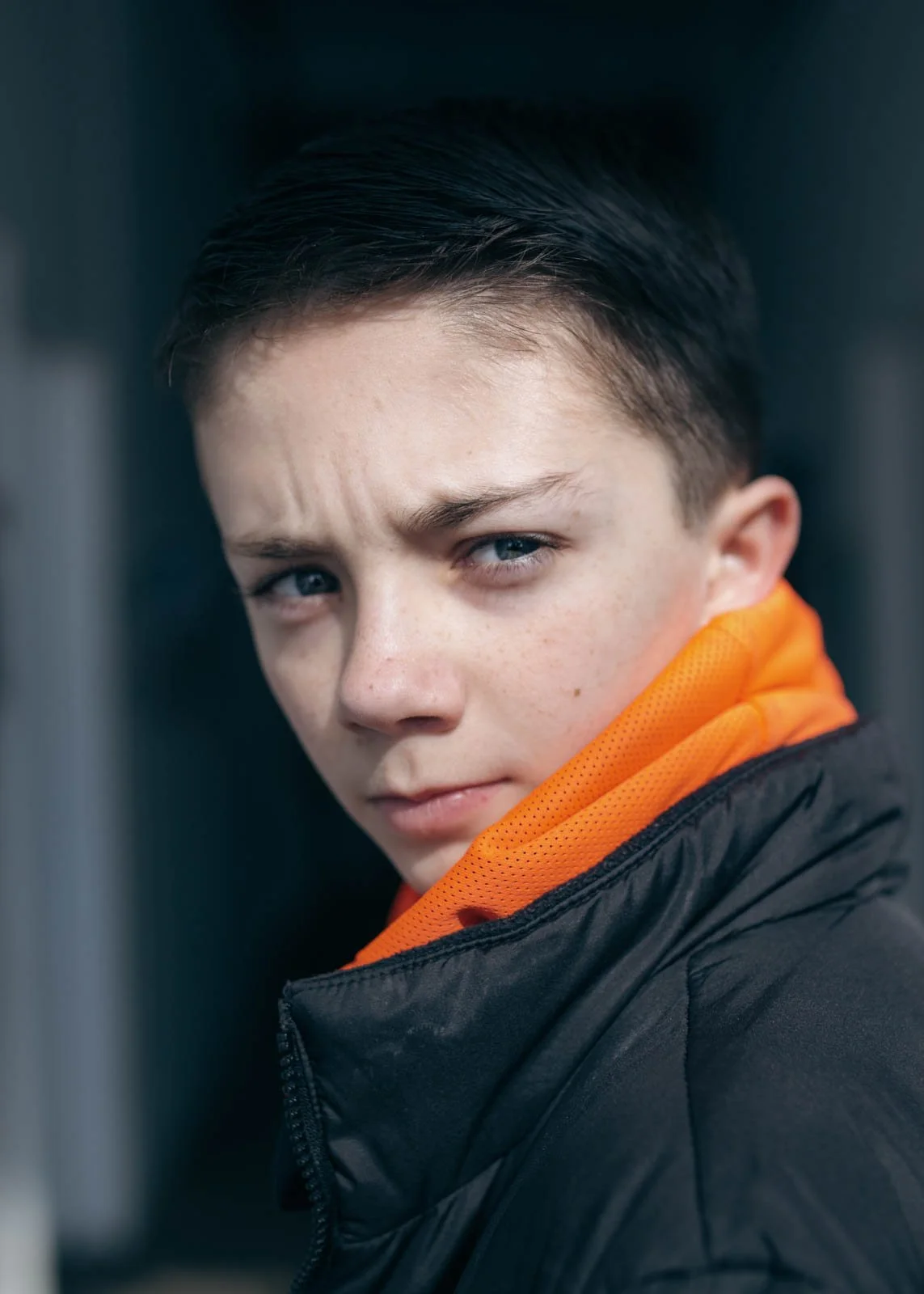 Close-up of a young boy with short dark hair, blue eyes, and freckles, wearing an orange hoodie and black jacket, gazing intently at the camera.
