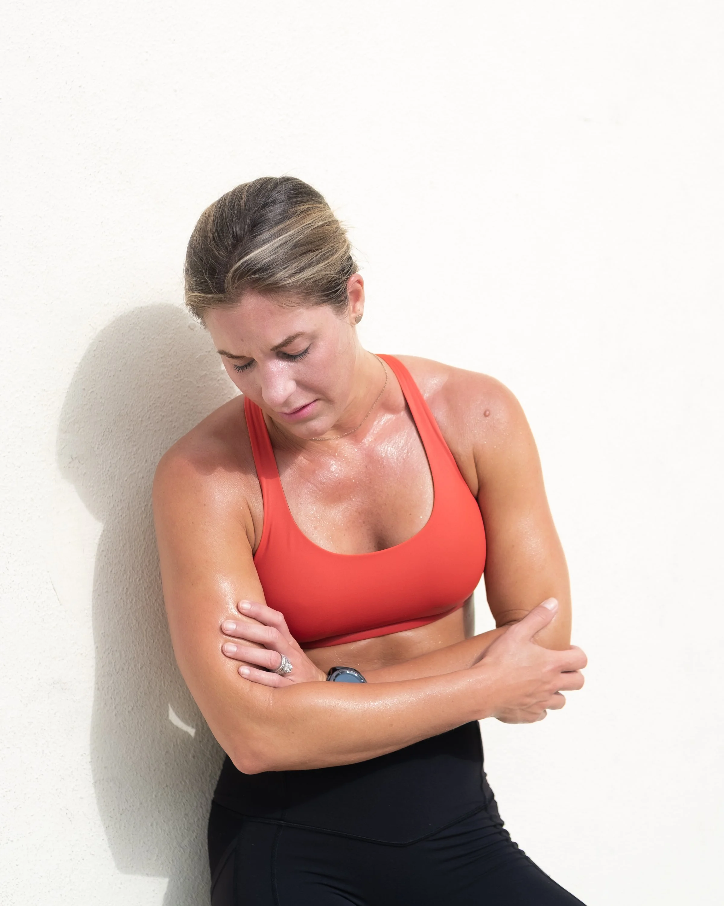 A woman in an orange sports bra and black leggings is stretching against a white wall, with her arms crossed and her head tilted down.