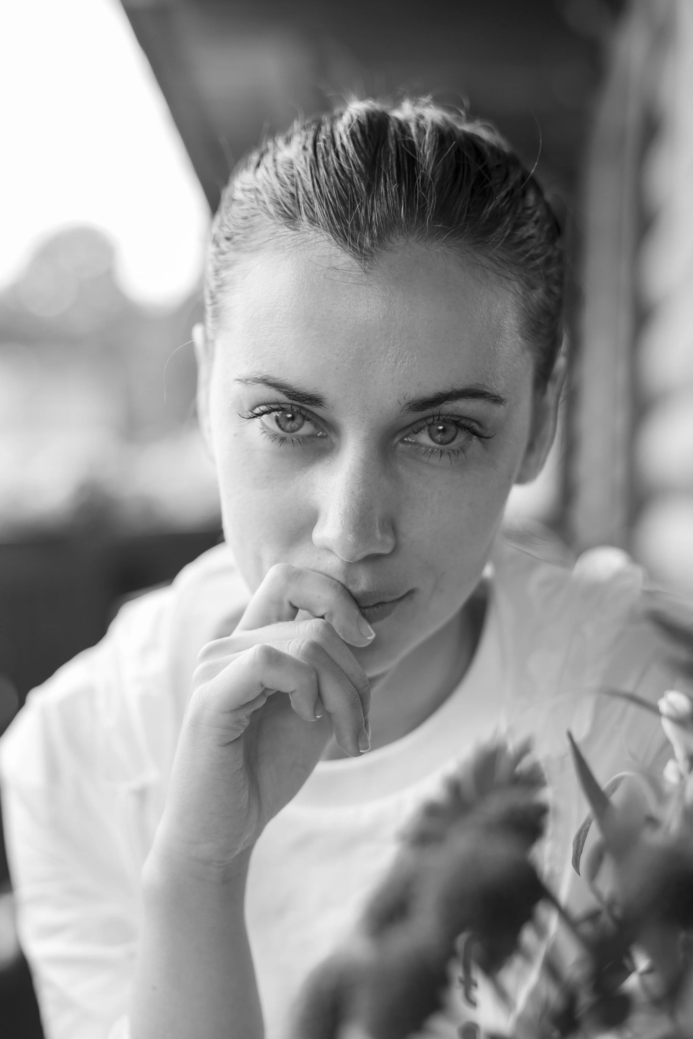 Close-up black and white photo of a young woman's face with focused eyes, hand near mouth, and her hair tied back.