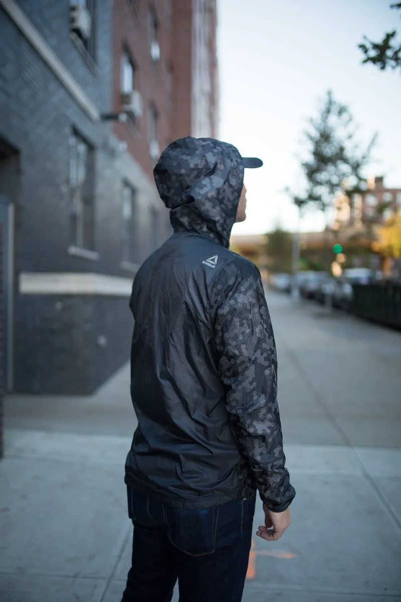 Person wearing a camouflage hooded Reebok jacket standing on a city sidewalk, seen from the back.