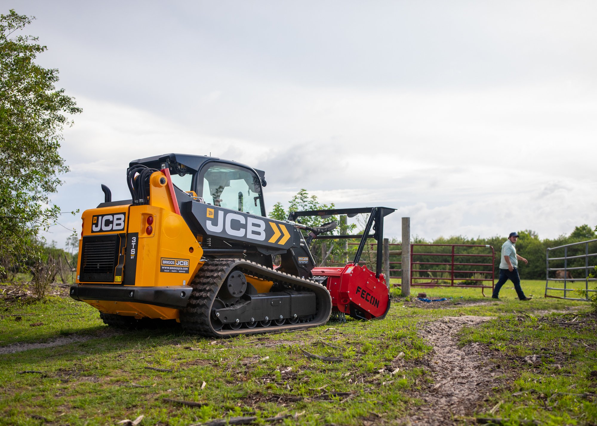 A small yellow and black JCB compact excavator working on a grassy field, with a man and a woman standing nearby and a cloudy sky overhead.