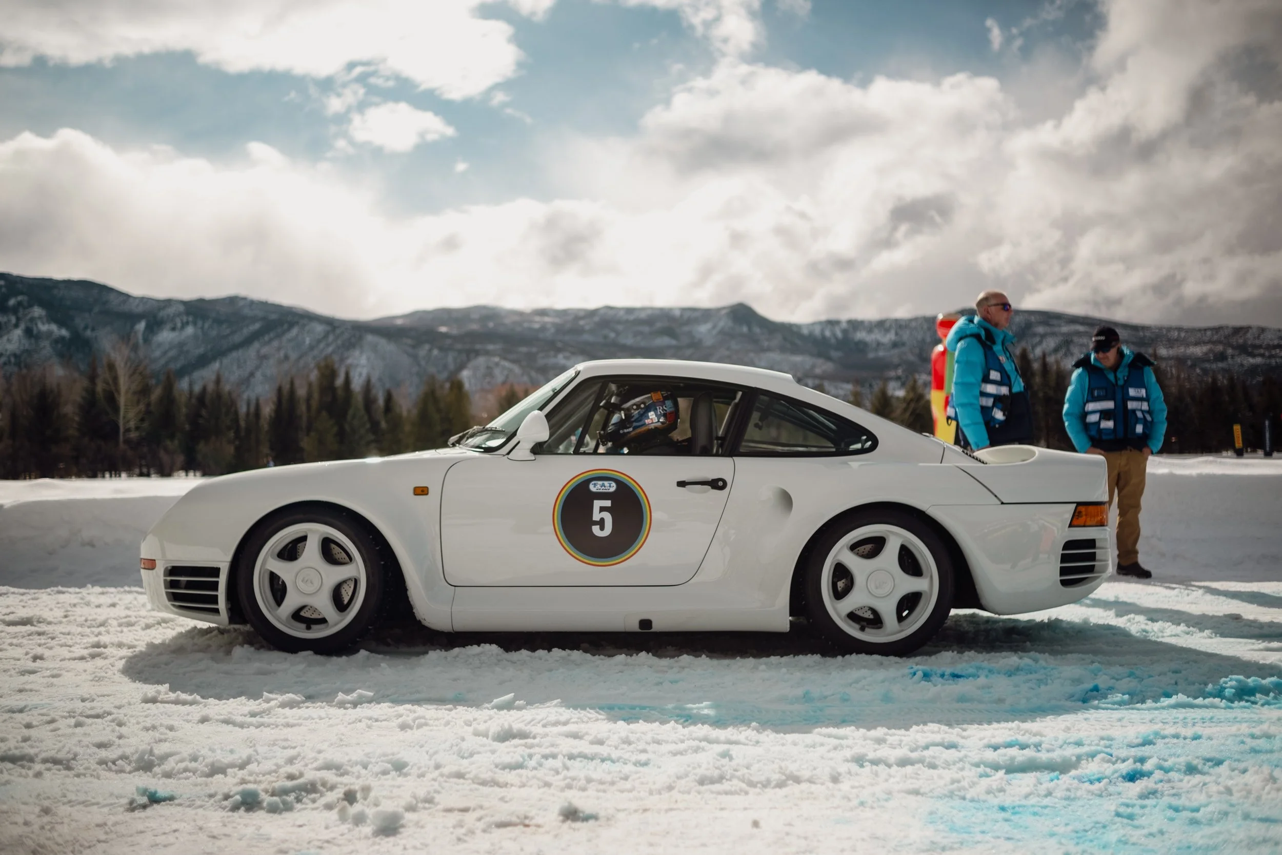 A white race car with racing decals parked on snow, with two men in blue jackets standing nearby, in a snowy landscape with mountains and clouds in the background. Porsche 959, Aspen Colordao