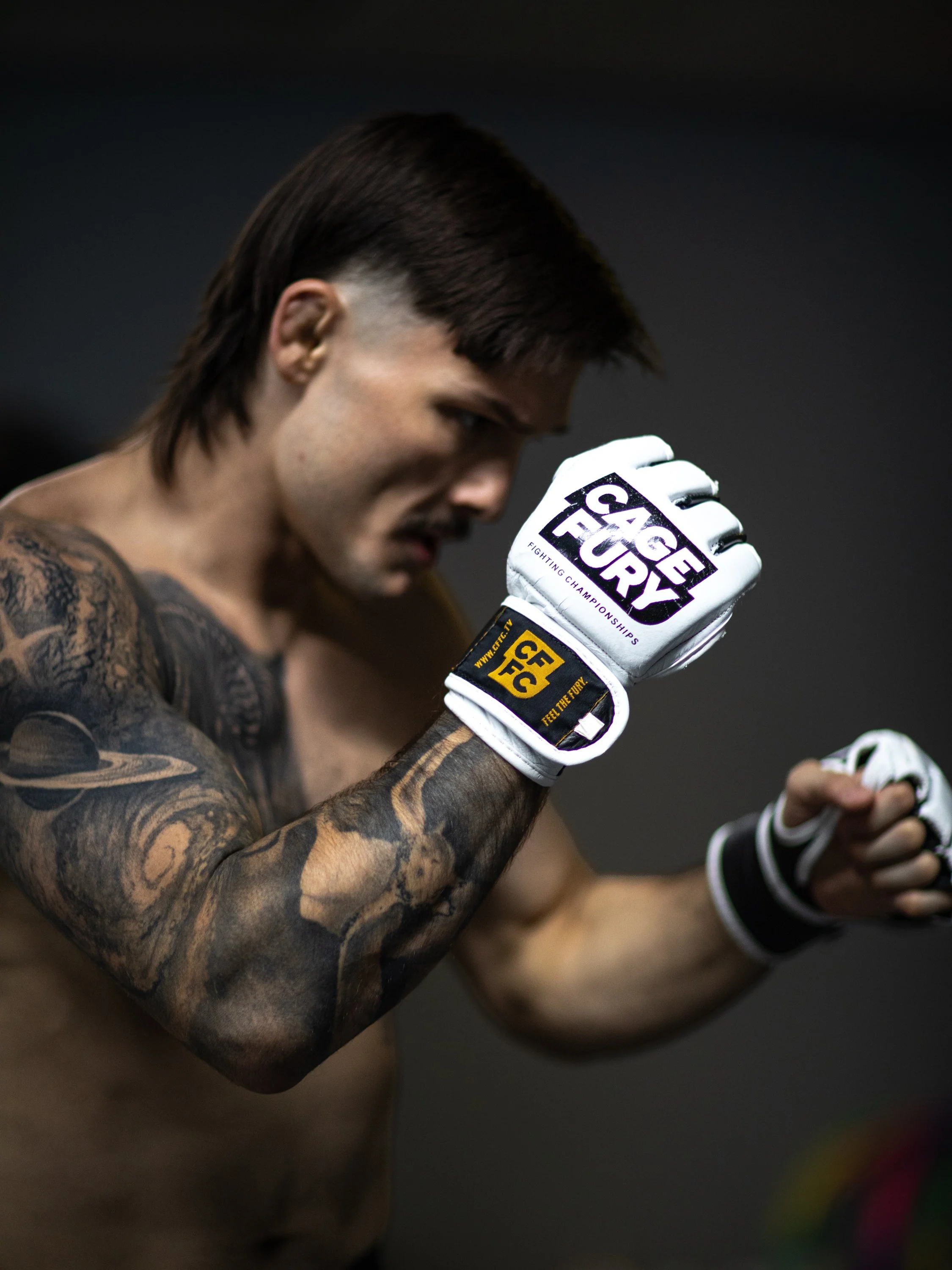 A male mixed martial arts fighter, shirtless with tattoos, wearing fight gloves, is preparing for a fight with his hands raised.