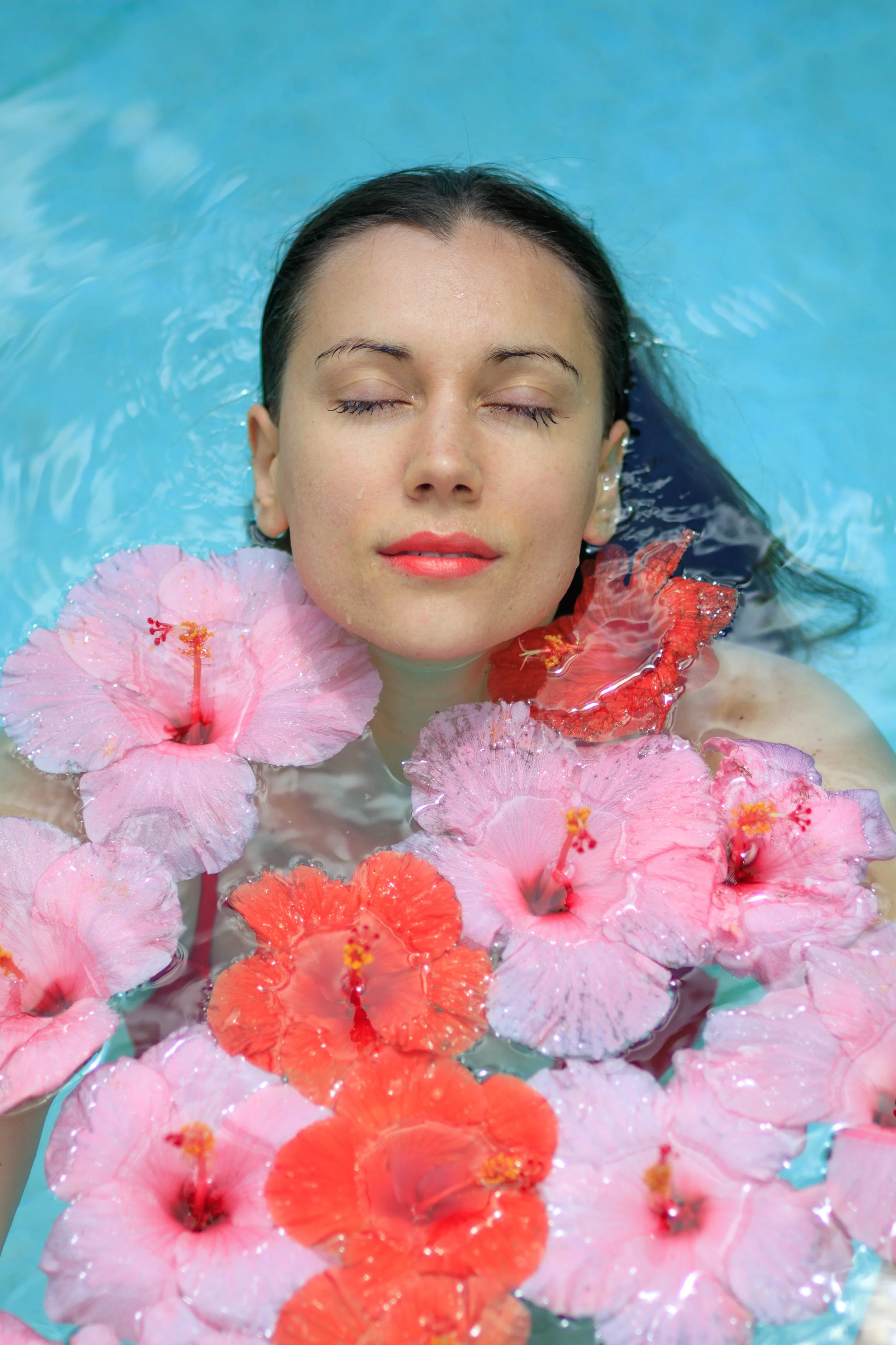 A woman with closed eyes floating in water, surrounded by pink and orange hibiscus flowers. Vogue italia.