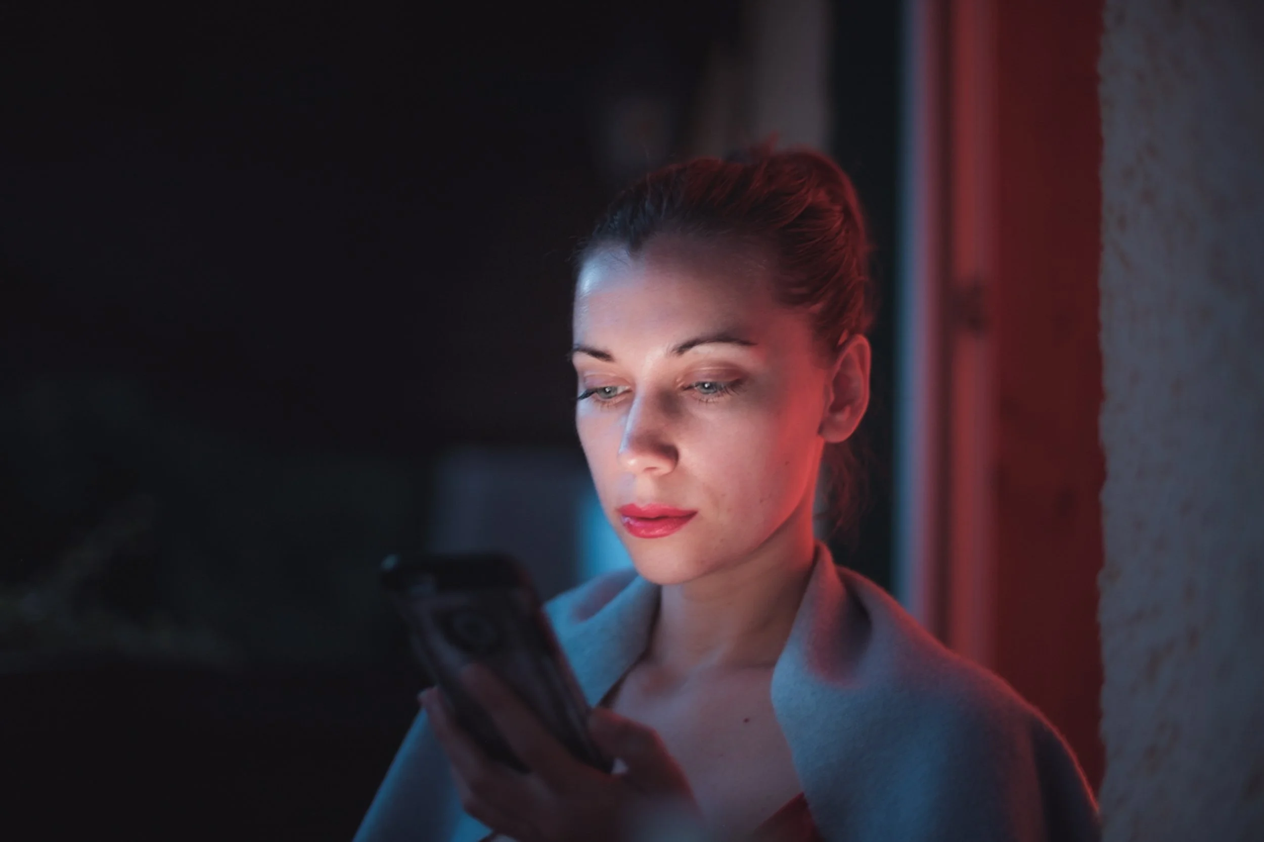 Close-up of a woman with red hair looking at her phone in a dimly lit room with red and blue lighting.