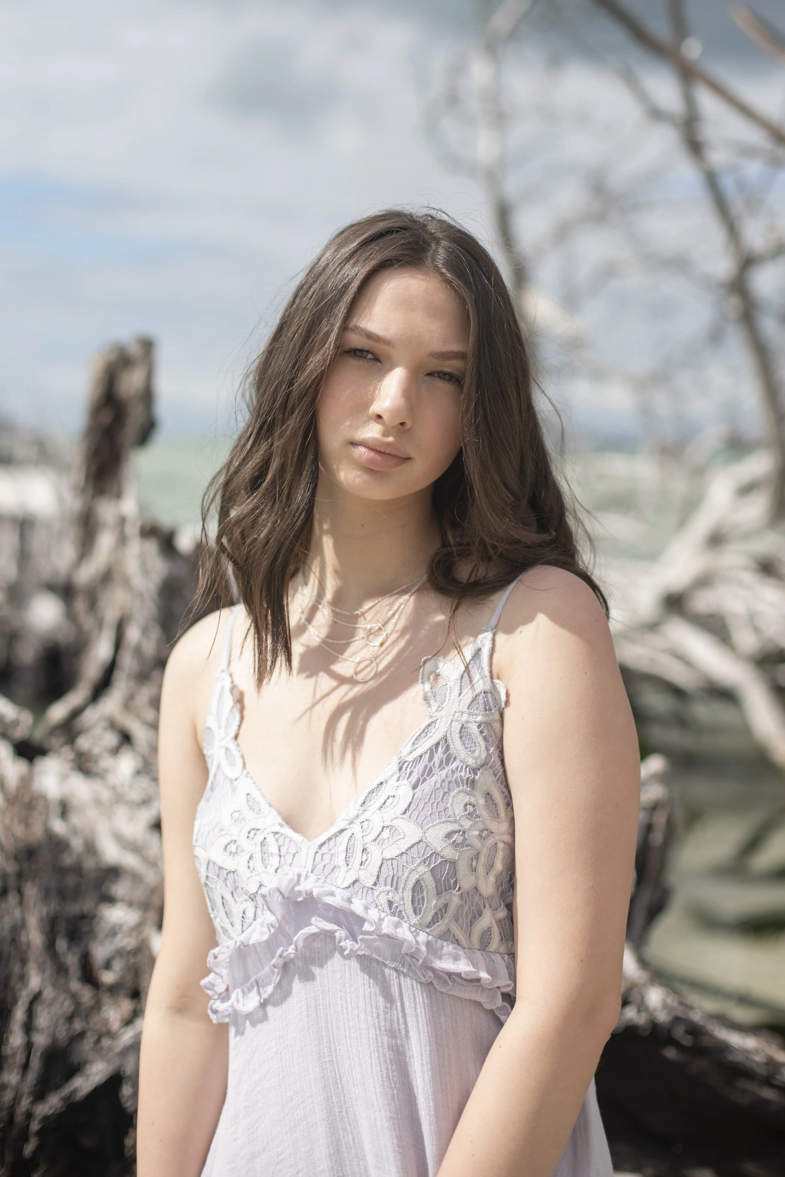 A young woman with long, wavy brown hair standing outdoors among driftwood and dried trees on a cloudy day, wearing a light-colored, lace dress.
