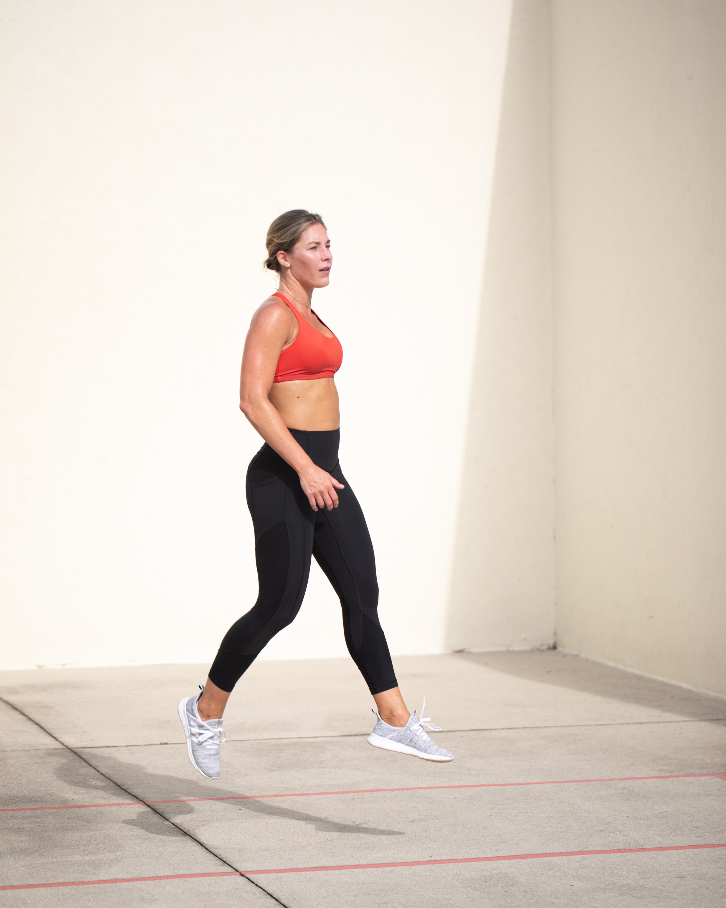 A woman in athletic clothing, including a red sports bra, black leggings, and gray running shoes, is captured mid-air during a jump on a concrete outdoor court with a plain beige wall background.