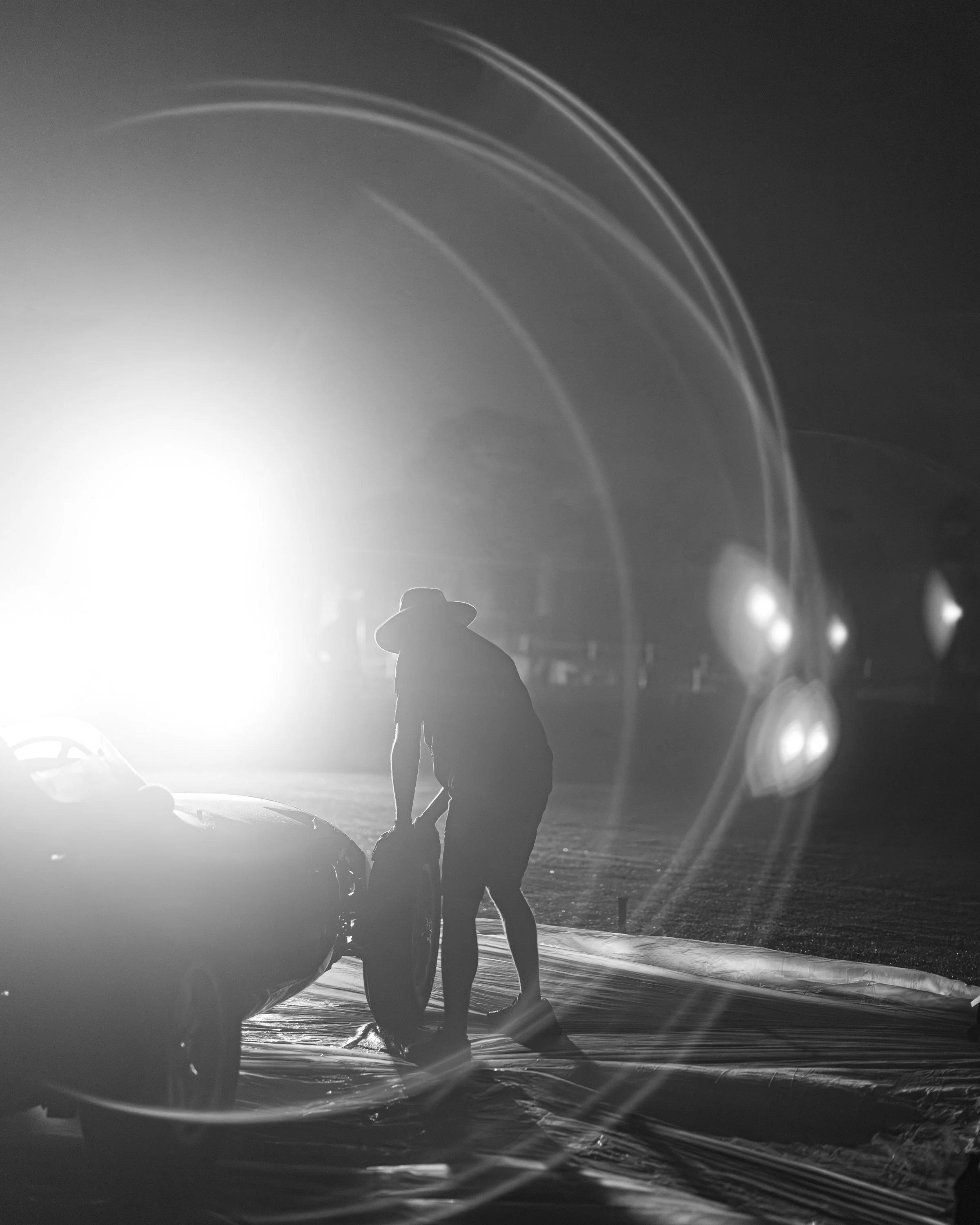 A black and white photo of a person holding an umbrella on the beach at night, standing next to a large beach ball, with wave patterns and light reflections creating a striped effect in the background.