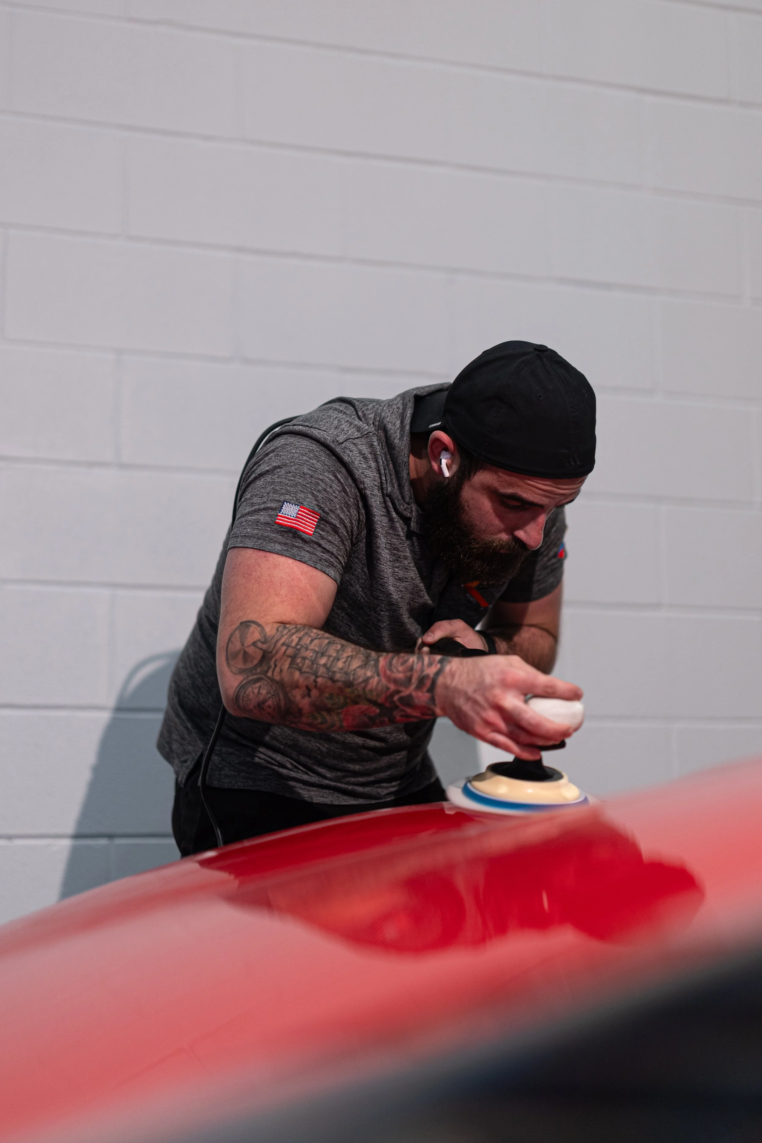 A man with tattoos, wearing a black cap and gray shirt with an American flag patch, is polishing a red car with a buffing machine inside a garage with plain white brick walls.