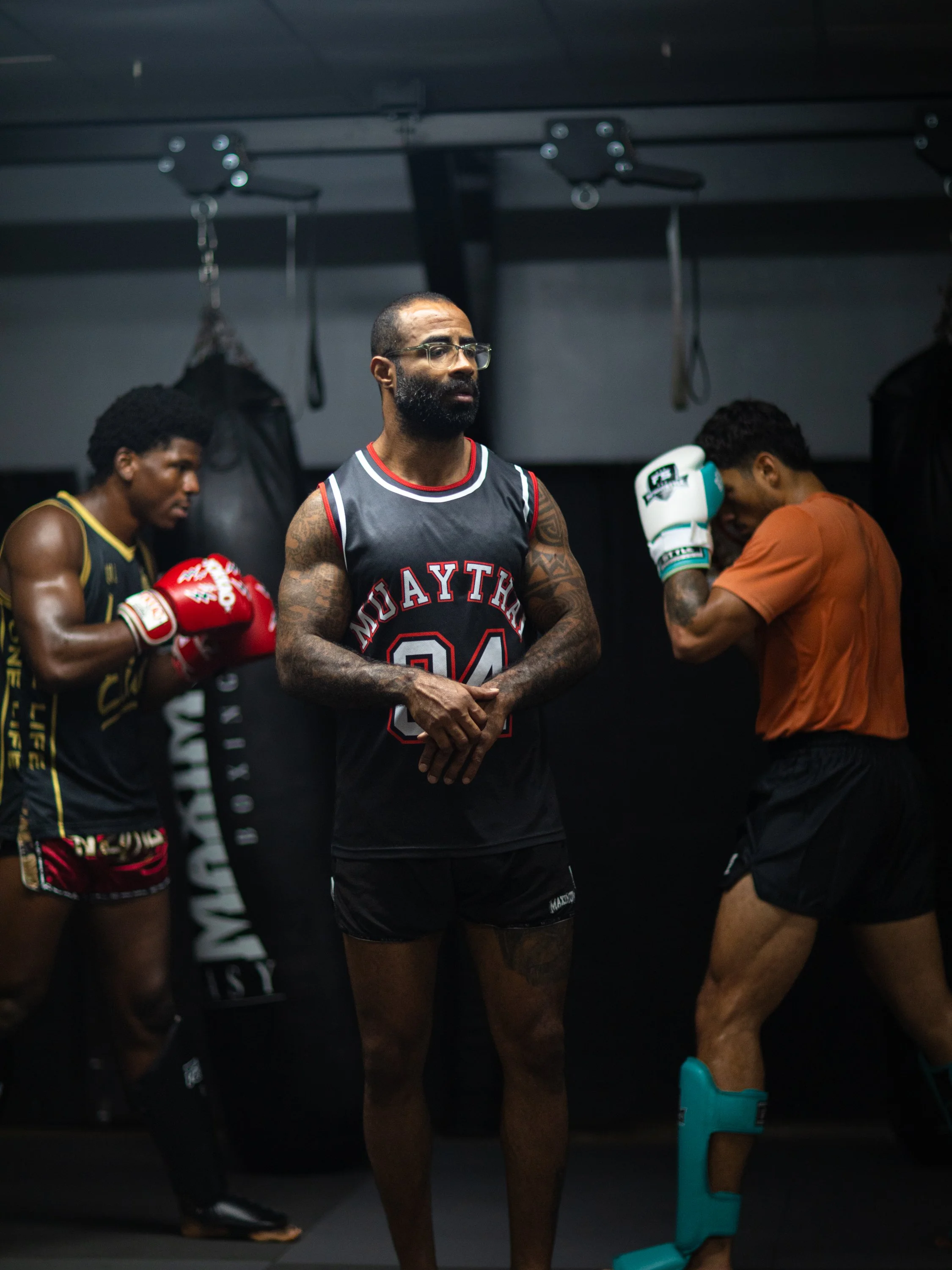 Three men in a gym, wearing Muay Thai shorts and gloves, standing with their heads bowed and eyes closed, possibly during a moment of silence or meditation.