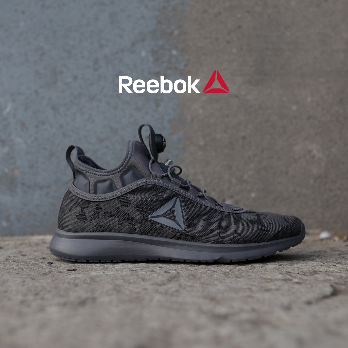 Black and gray Reebok athletic shoe on a concrete surface with a concrete wall background, featuring the Reebok logo on the side. Reebok.
