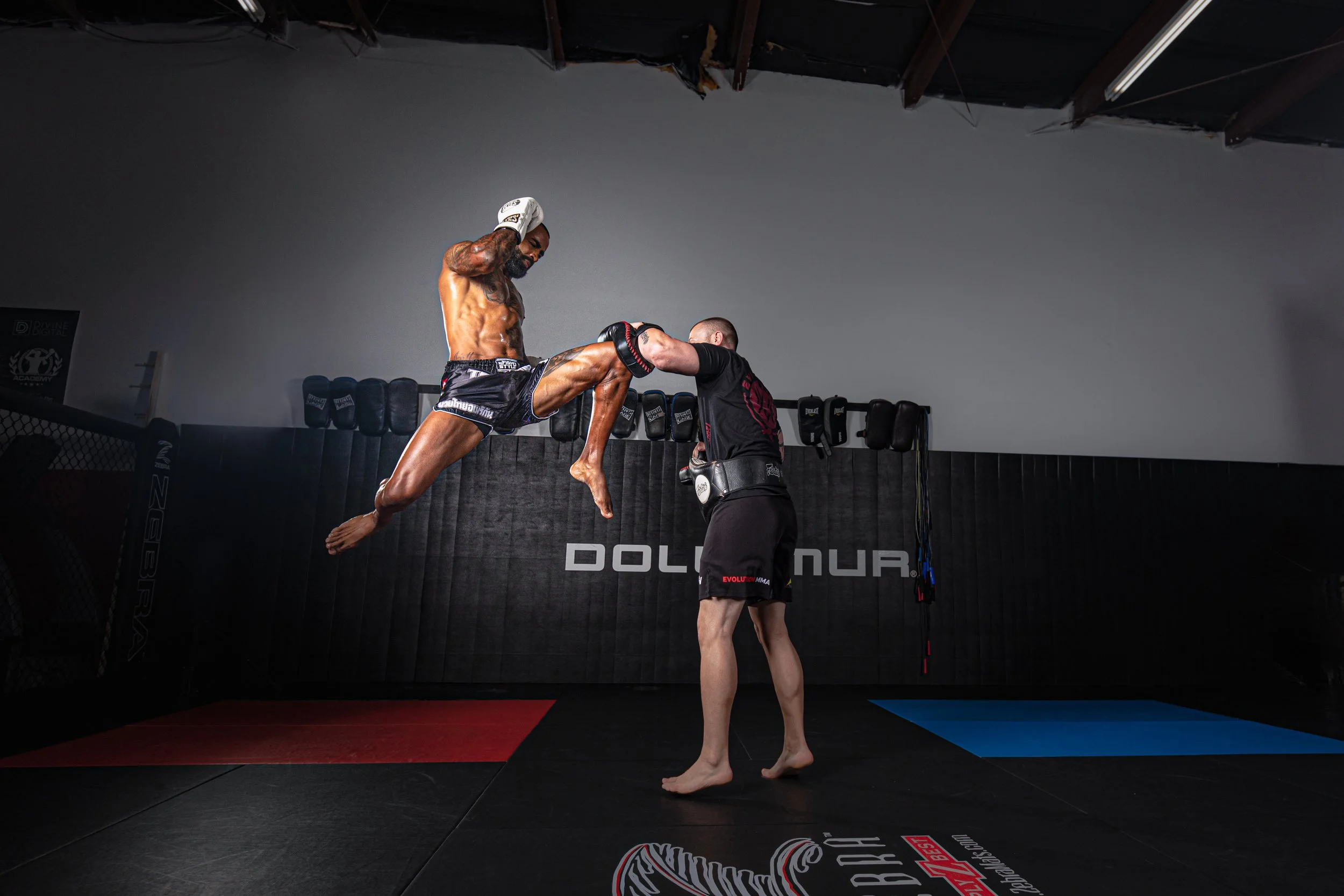 A shirtless man with tattoos on his arms and chest practicing a martial arts move with a trainer in a gym. The man is mid-air, executing a kick towards the trainer, who is holding pads. The gym has black padded walls and equipment in the background.