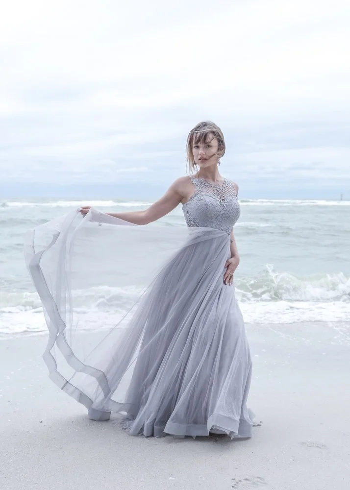 A woman in a flowing silver-gray gown standing on a beach with ocean waves in the background.