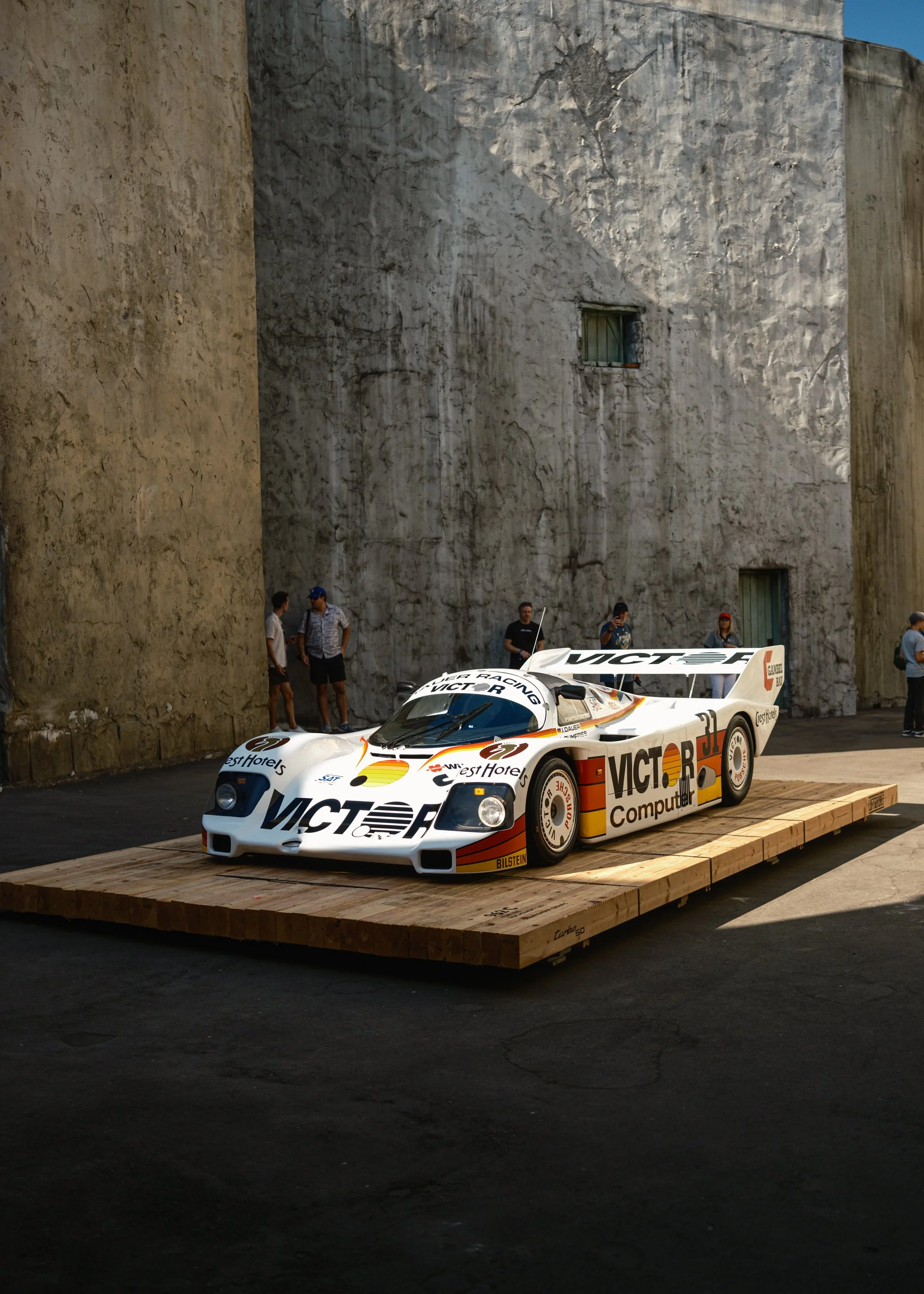 A white race car with orange and black accents, bearing various sponsor logos, is on a wooden platform in front of a large textured concrete wall. Several people are standing nearby, discussing and taking photos of the car.