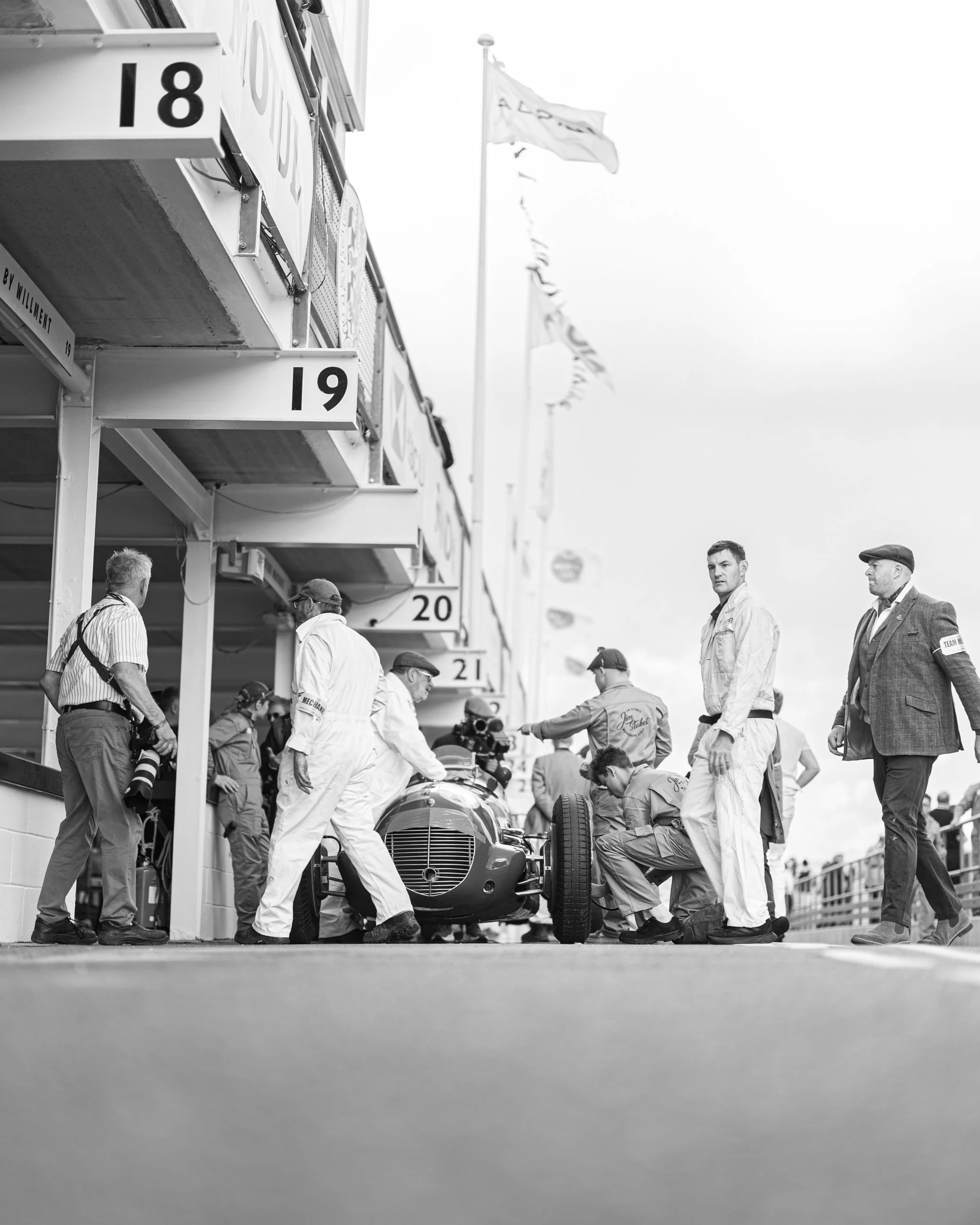 A vintage race car being serviced by a team of mechanics on a race track. The car is aligned with the pit lane, and several people are gathered around it, some kneeling and others standing, with equipment and tools.