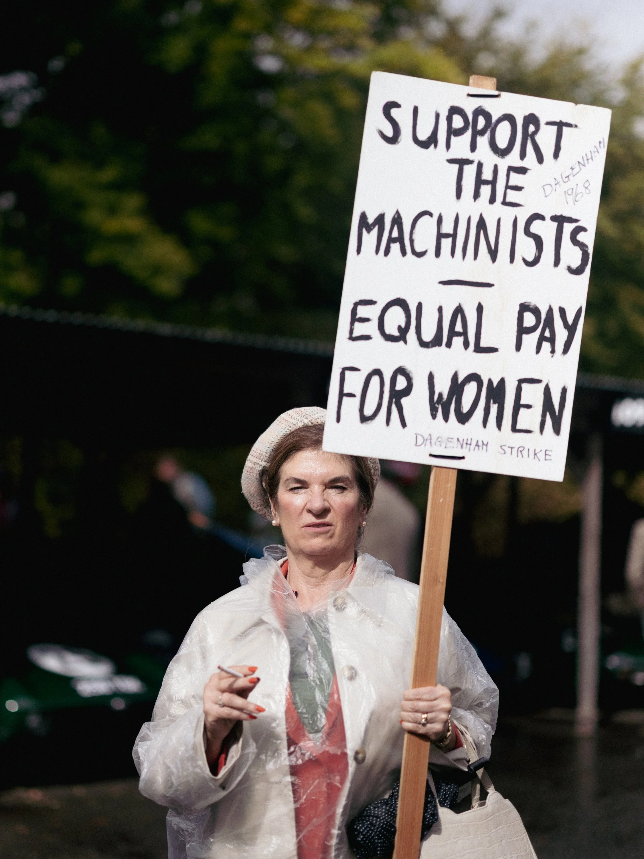A woman participating in a protest holding a sign that reads support the machists equal pay for women. She is wearing a hat, a transparent plastic coat, and has a serious expression on her face. The background is blurred with trees and other people.