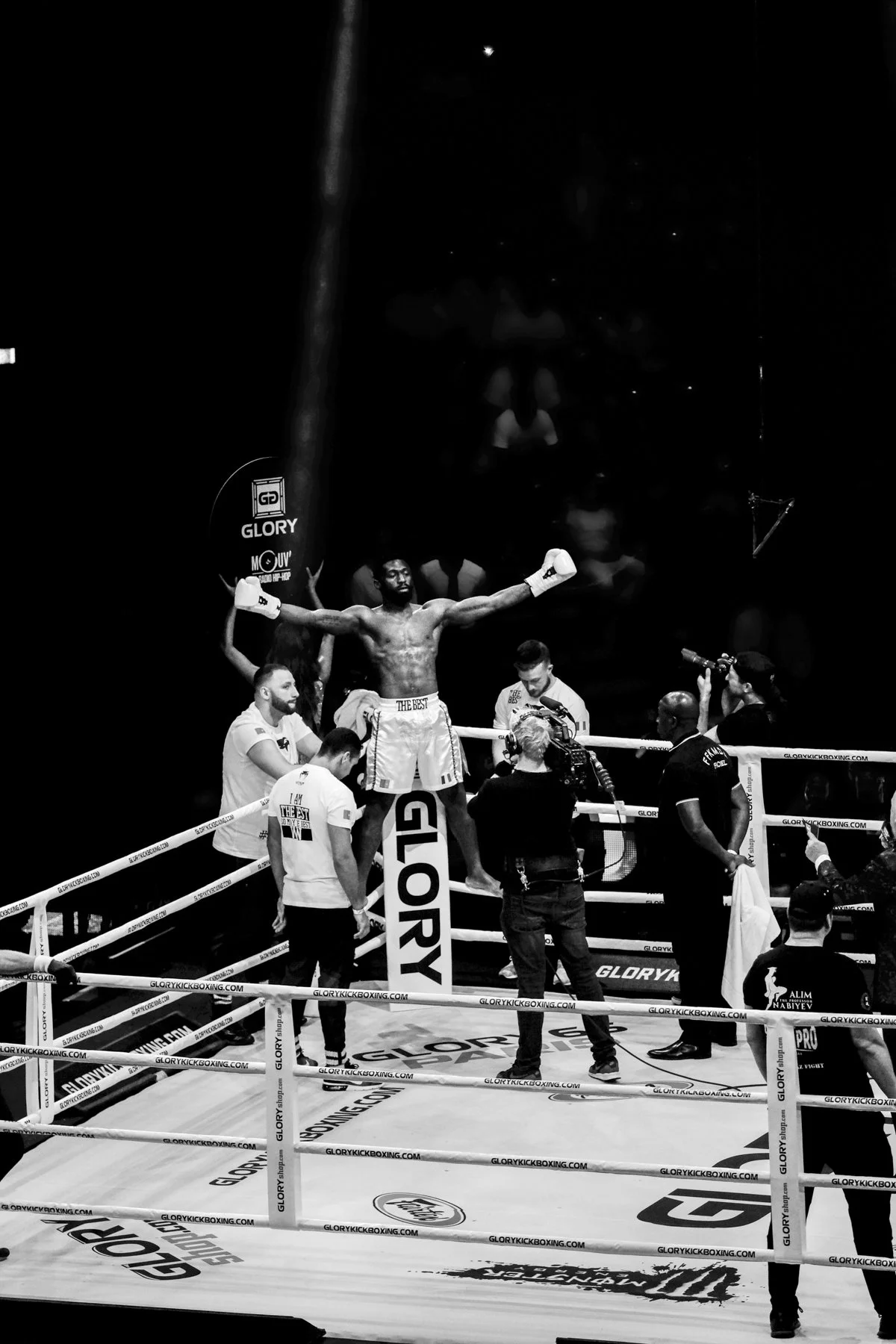 A victorious male boxer with outstretched arms in a boxing ring, surrounded by officials, photographers, and spectators, celebrating after a match. The scene is in black and white.