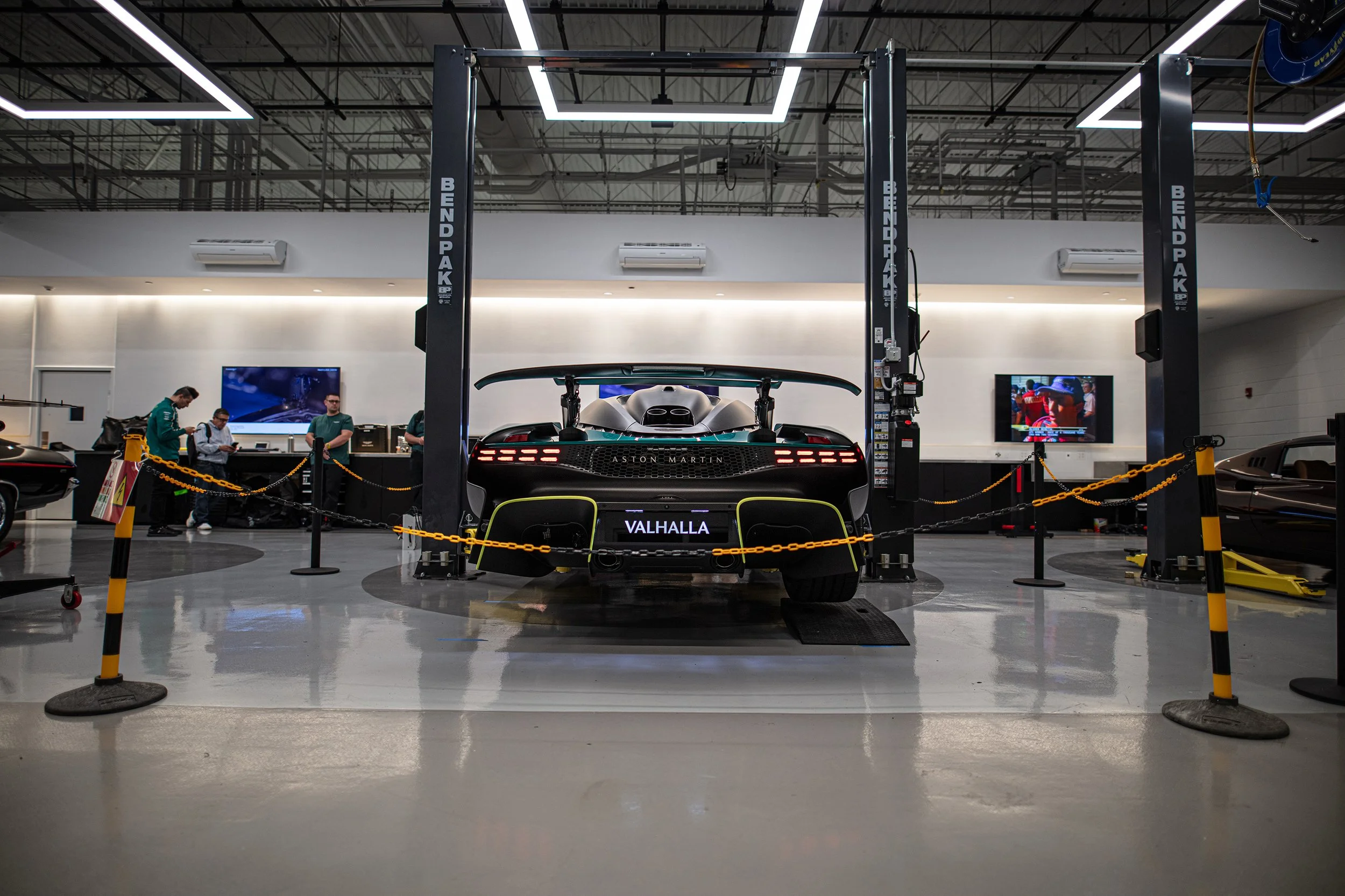 A black Aston Martin Valhalla sports car displayed inside a modern showroom, surrounded by yellow chains, within an industrial-style space with exposed ceiling pipes and large TV screens on the wall.