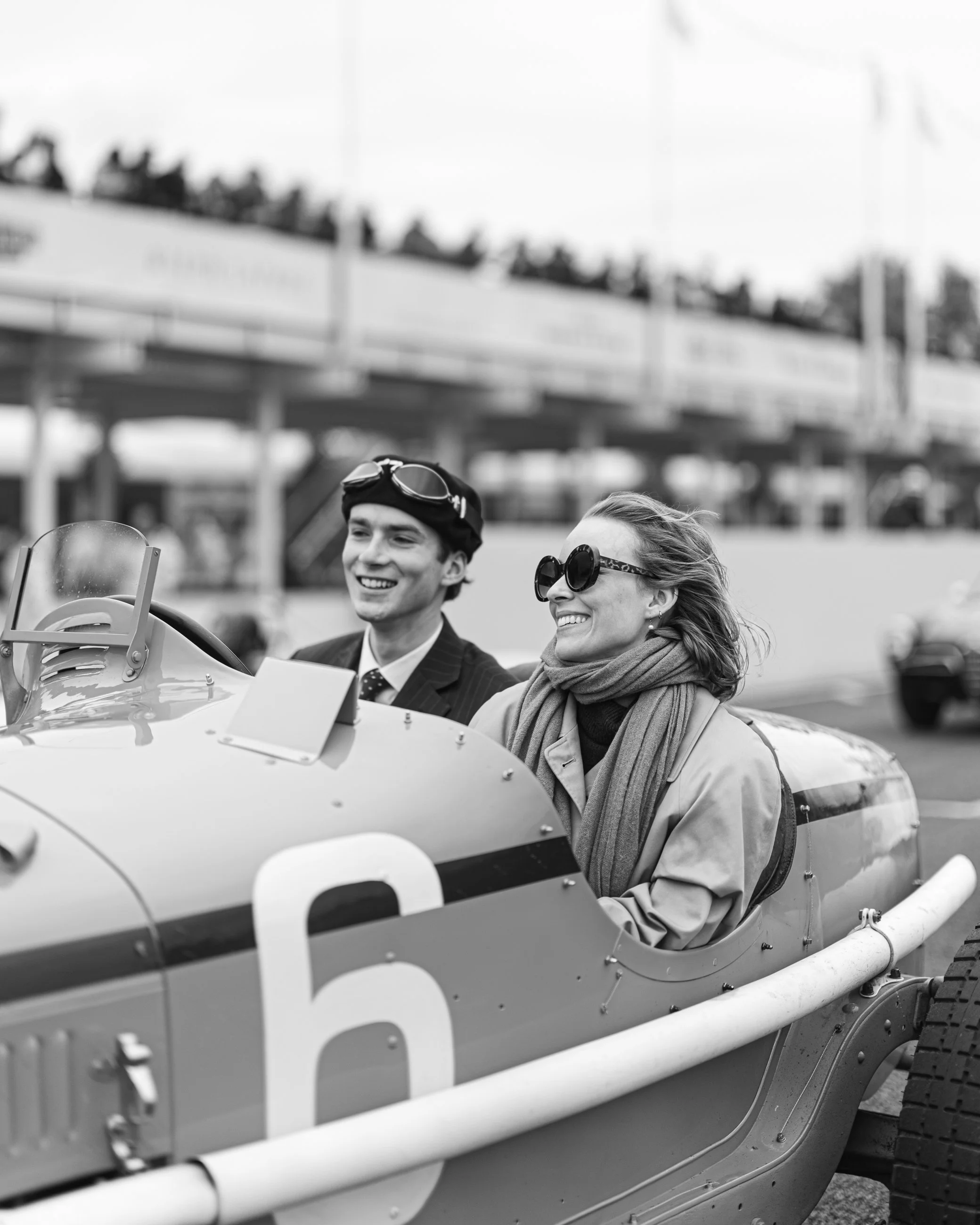 Black and white photo of two smiling women sitting in a vintage race car, one wearing goggles and a cap, the other with sunglasses and a scarf. A group of people is visible on an elevated structure in the background.