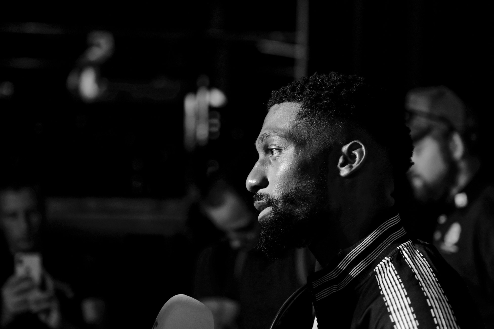 A black and white photograph of a man with short curly hair and a beard, dressed in a striped blazer, sitting indoors with blurred people in the background. Cedric Doumbe