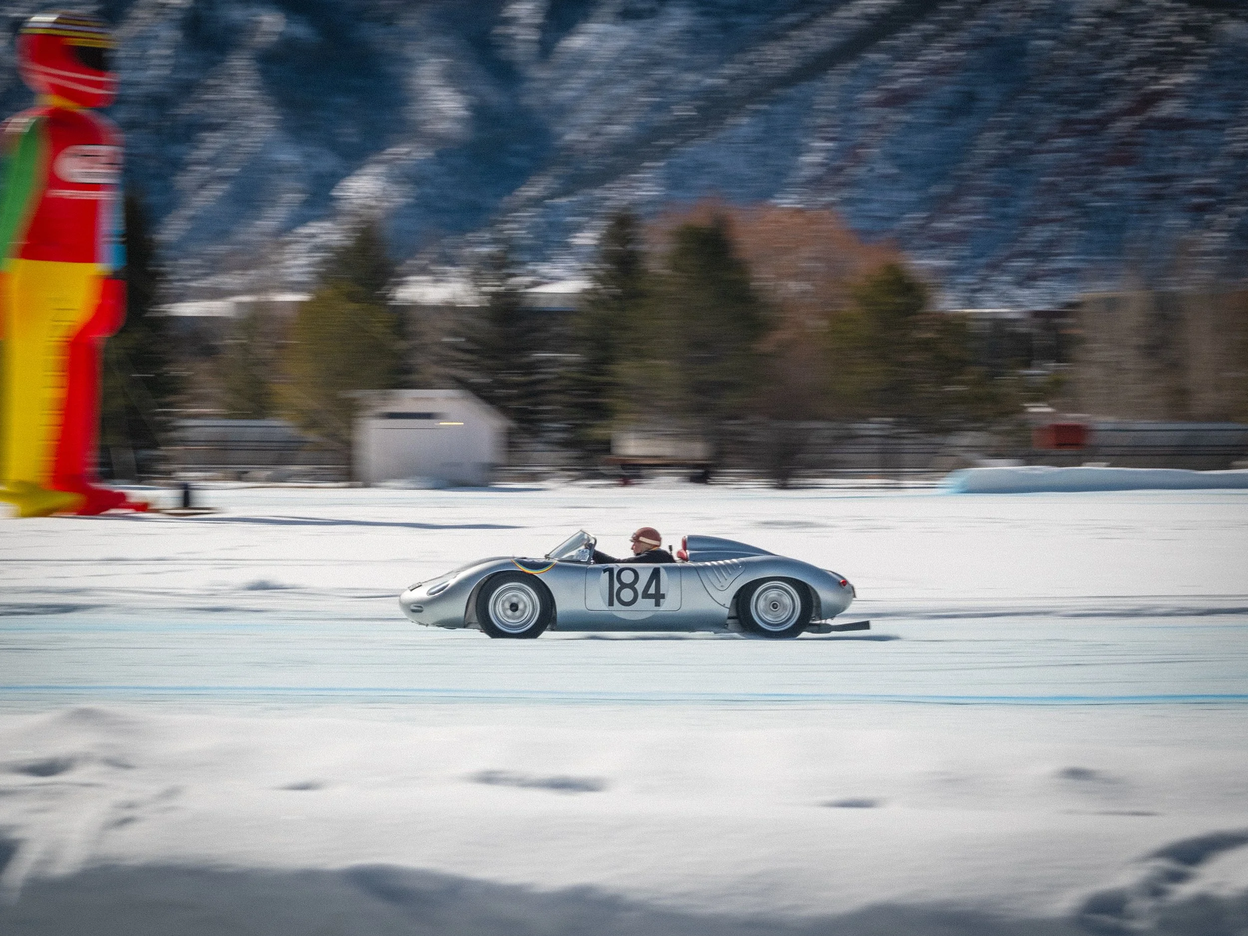 A vintage silver race car with number 184 on its side, speeding on a snowy mountain landscape with snow-covered trees and mountains in the background. Porsche race car, Jeff Zwart, Fat Ice Race, Aspen Colorado