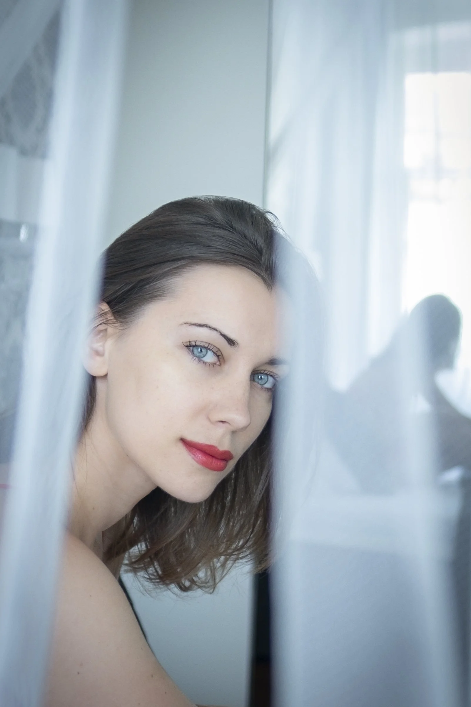 A woman with blue eyes, dark brown hair, and red lipstick looking over a sheer curtain with a soft light background.