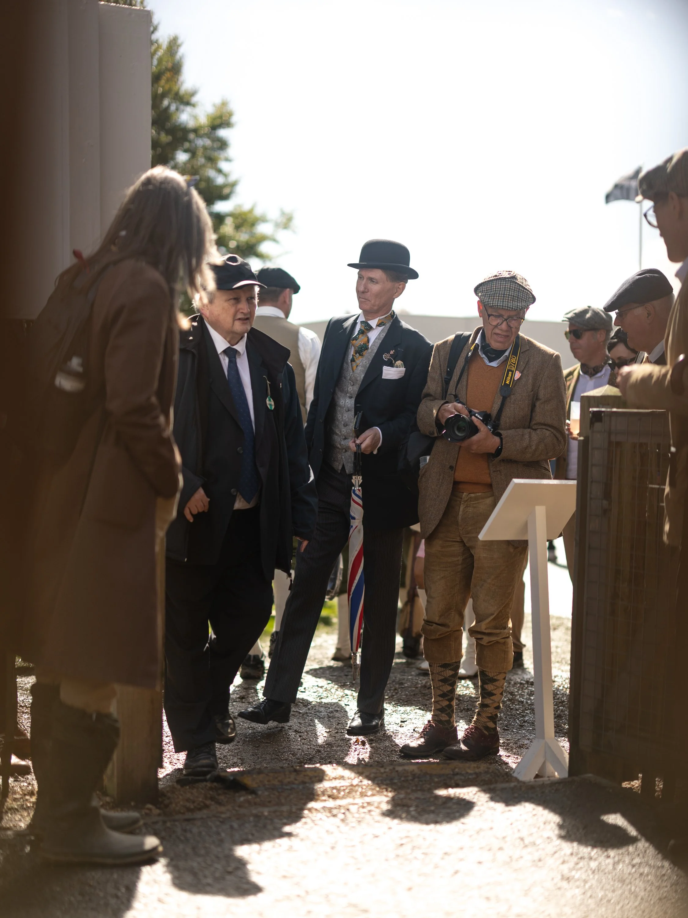 A group of men dressed in vintage or early 20th-century attire gathered outdoors, engaged in conversation, with some holding cameras, and a bright sky in the background.