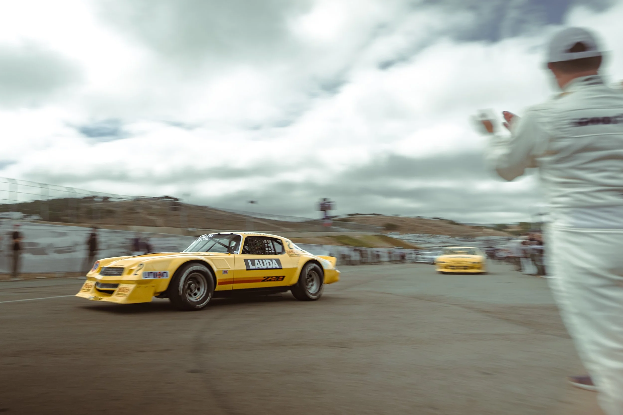 A yellow race car with the word 'LAUDA' and the number '028' on it driving on a track, with racing personnel in white uniforms and headphones nearby, and a cloudy sky overhead. Patrick Long