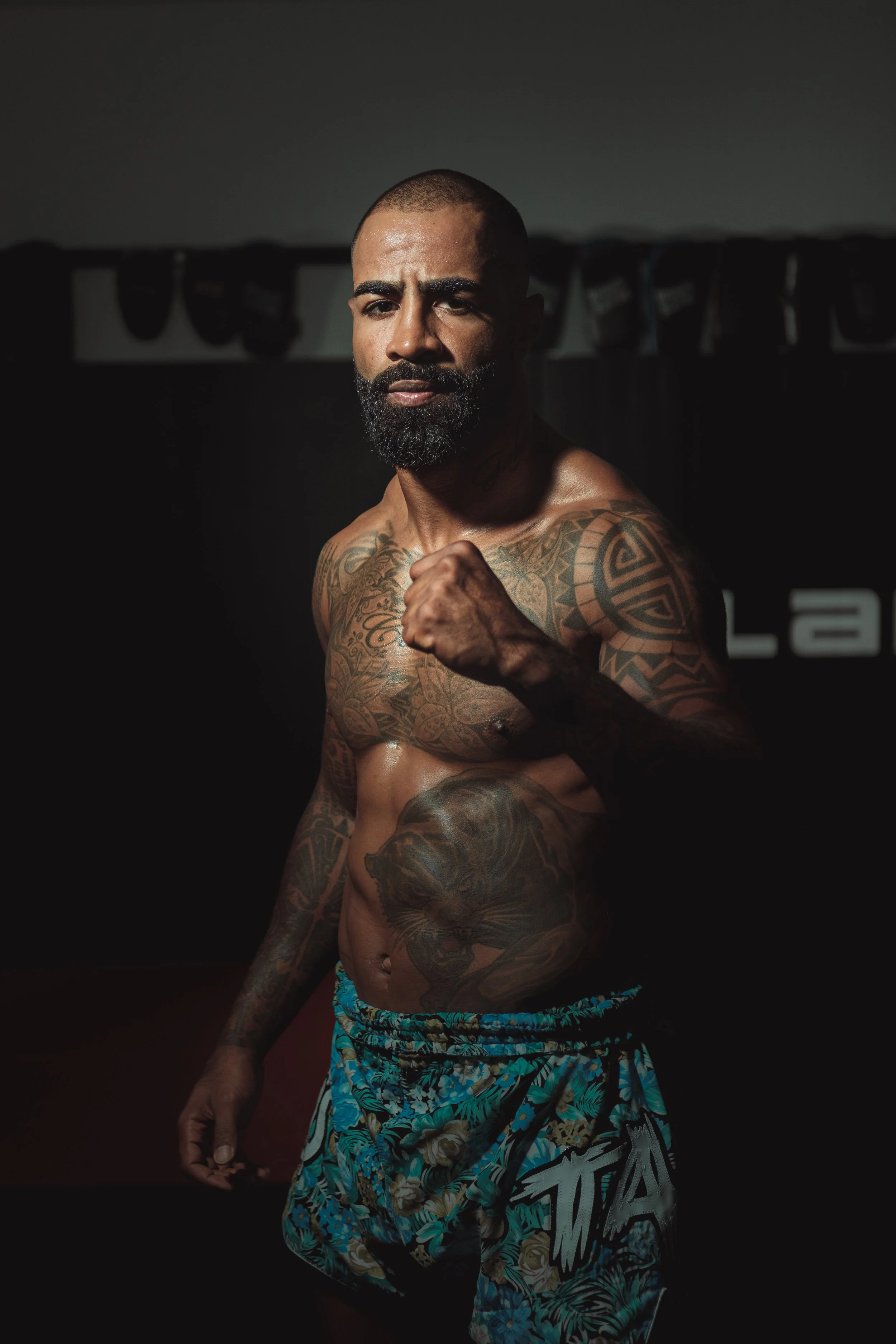 A muscular man with a beard and tattoos posing shirtless in front of a dark background. He is wearing colorful floral shorts and making a fist with his right hand. Cosmo Alexandre