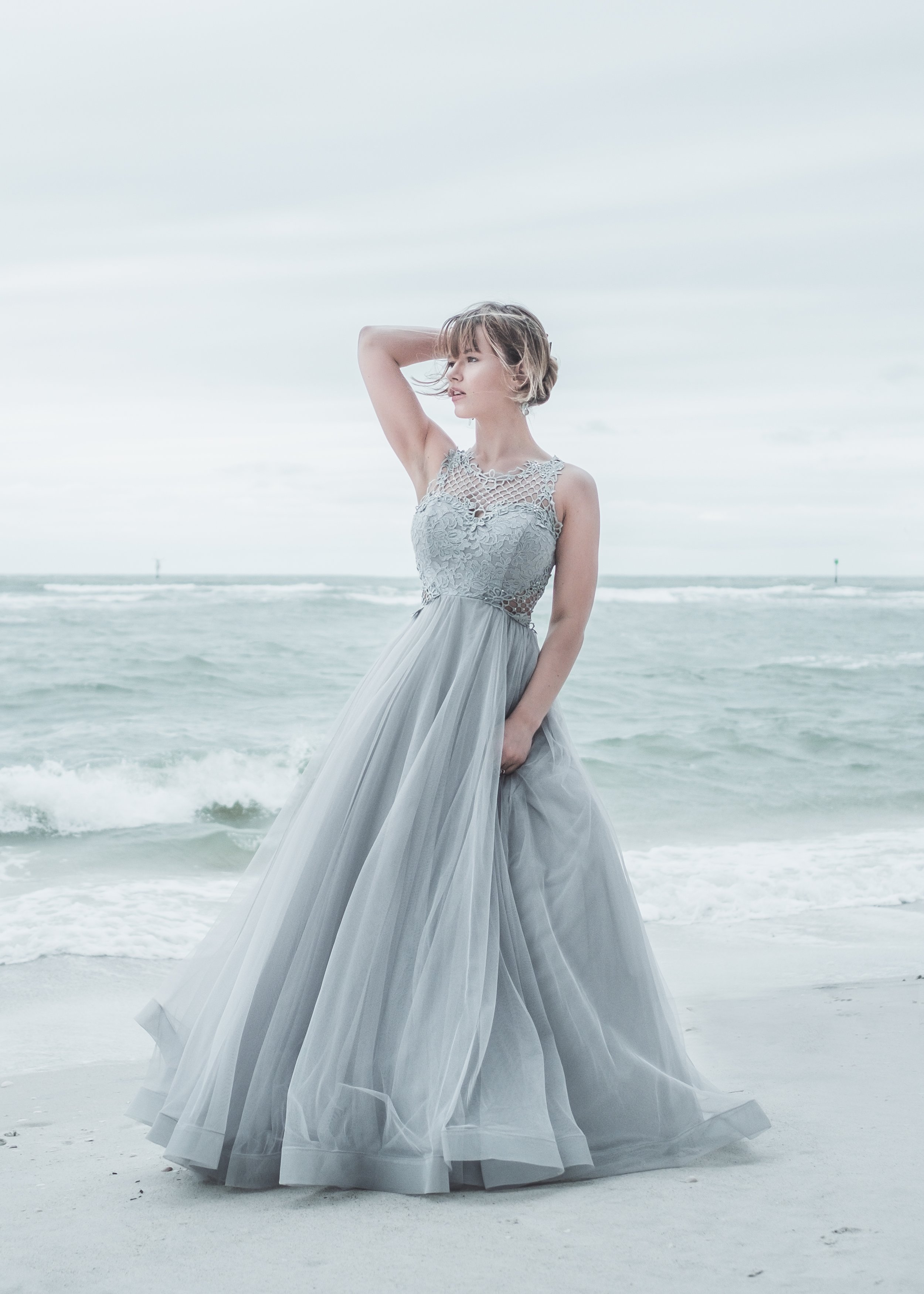 A woman in a flowing, intricate dress standing on the beach with ocean waves behind her, looking to the side with her hand in her hair.