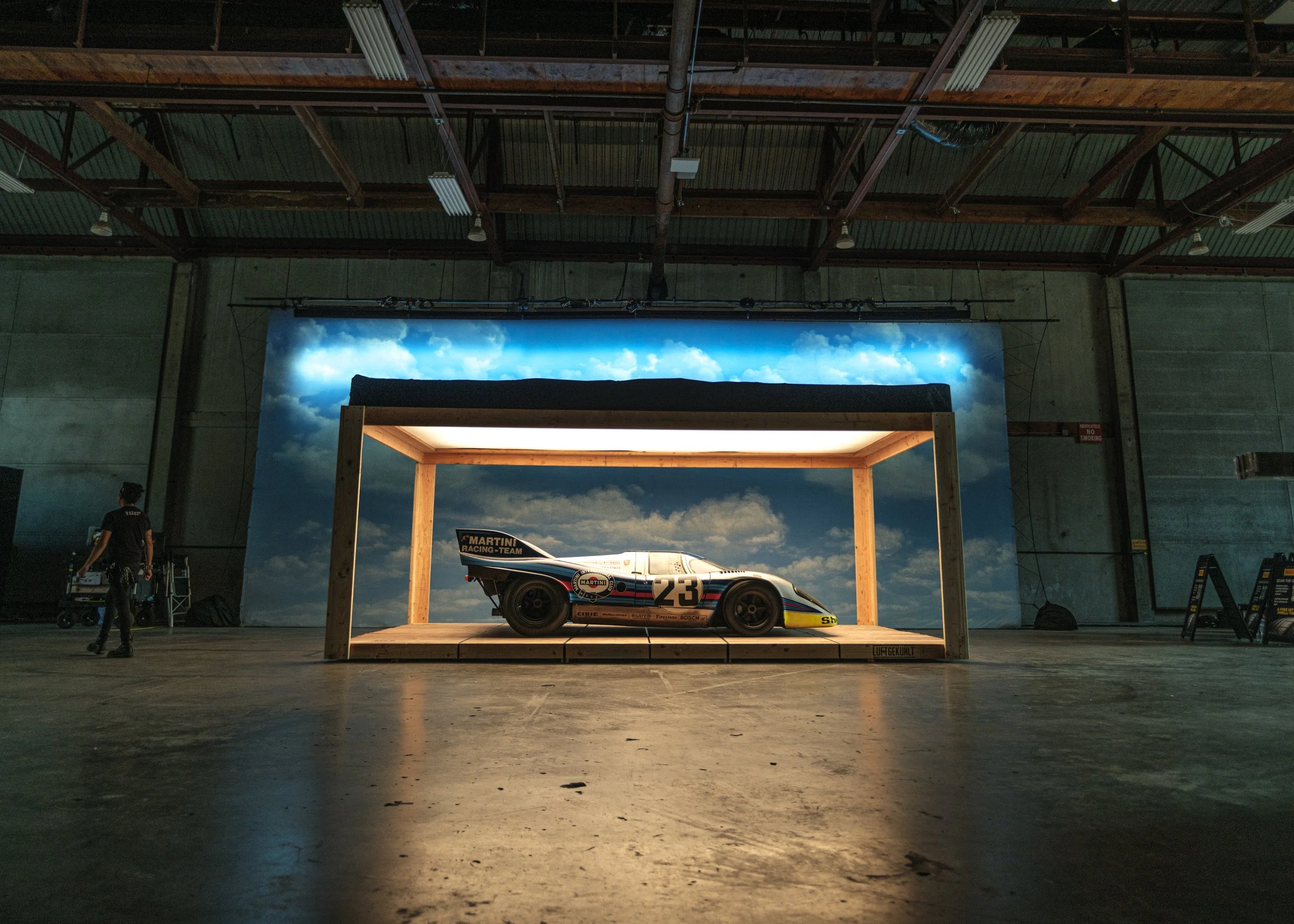 Race car displayed on a wooden platform inside a large industrial warehouse with a painted sky backdrop. Luft.