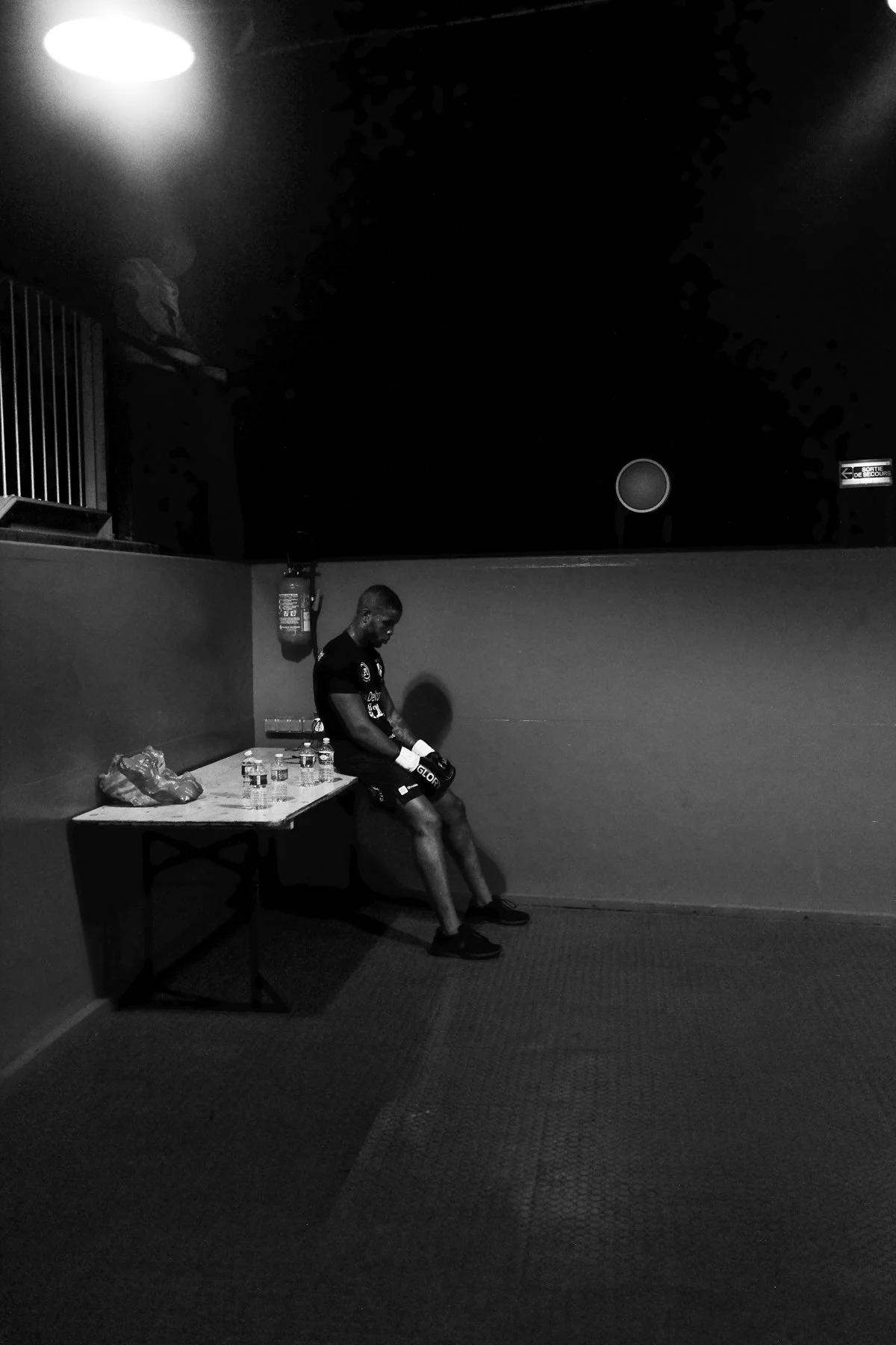 A lone athlete sits on a bench in a dimly lit room, wearing boxing gear, with water bottles and a plastic bag on a nearby table.